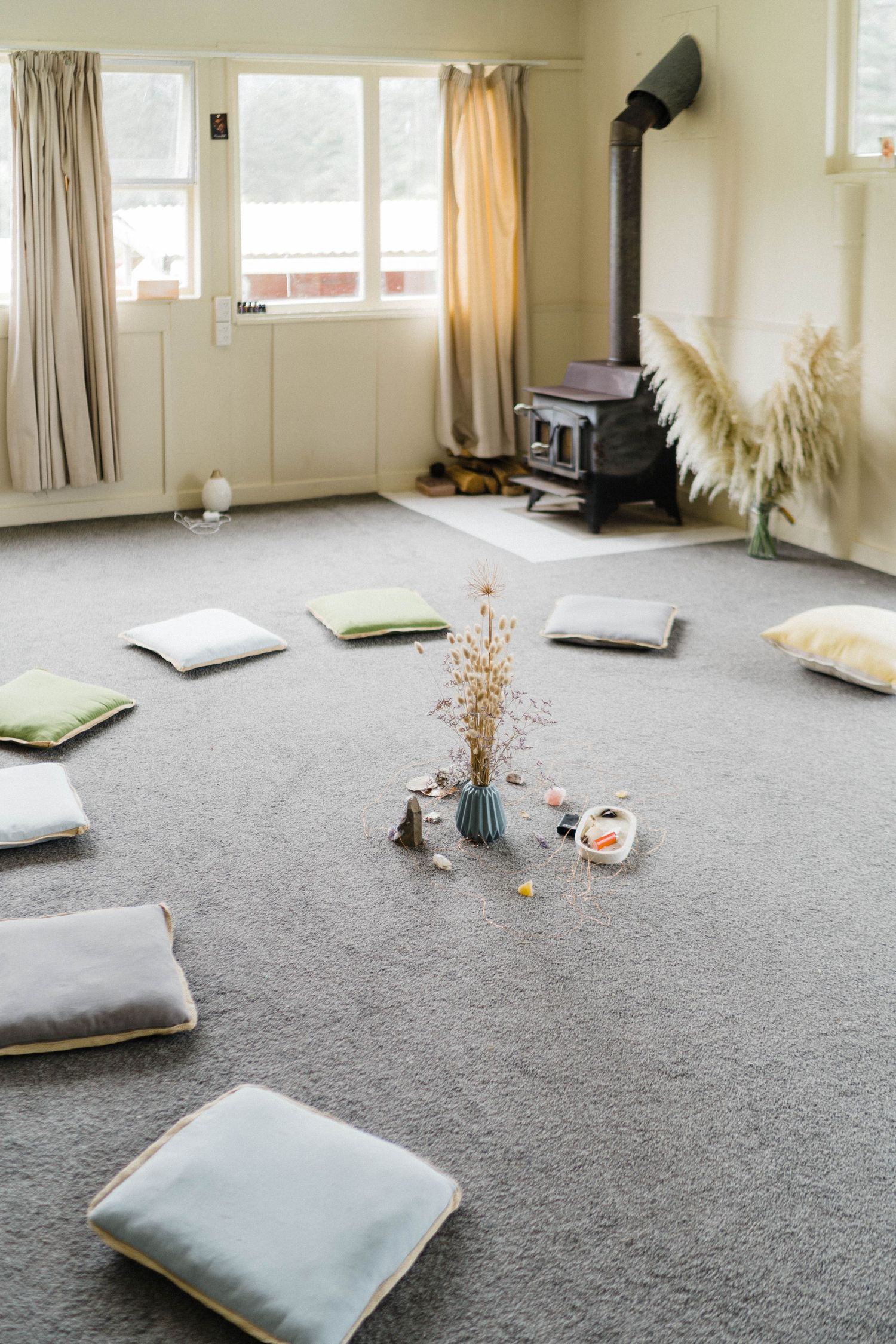 A cozy room with floor cushions arranged in a circle around dried flowers, featuring a wood stove in the corner.