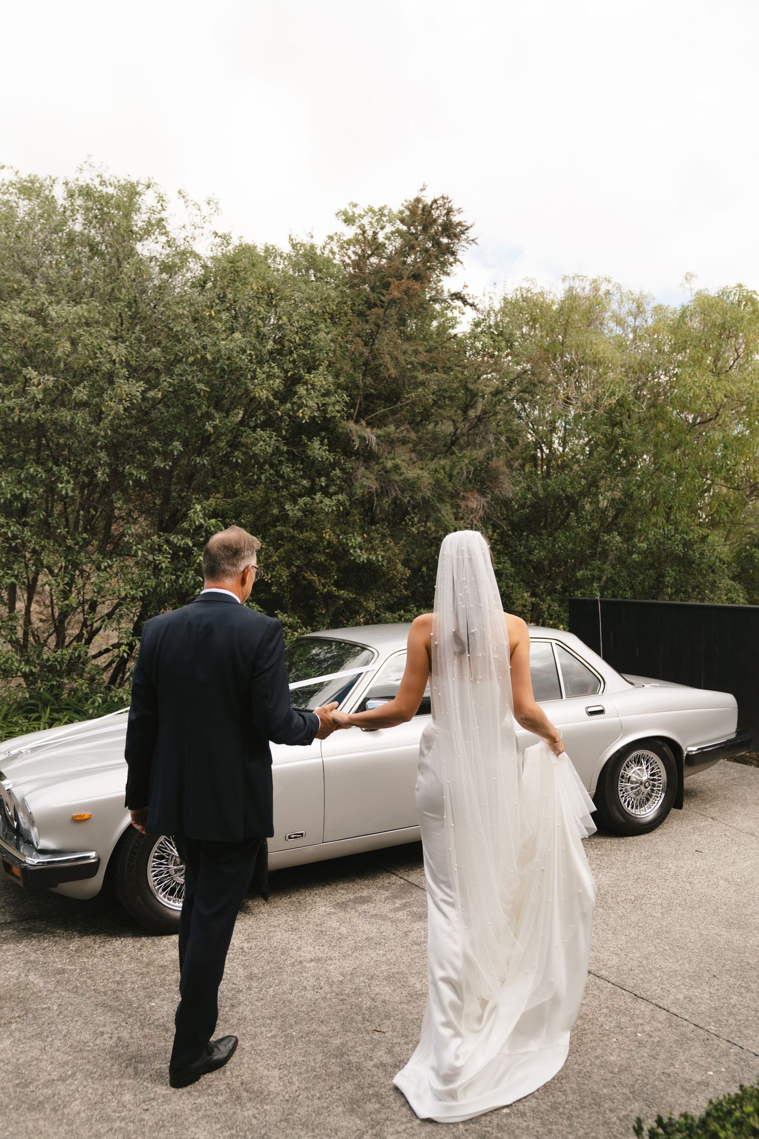 Bride in flowing white gown and groom in black suit approach a classic white vintage car on their wedding day.