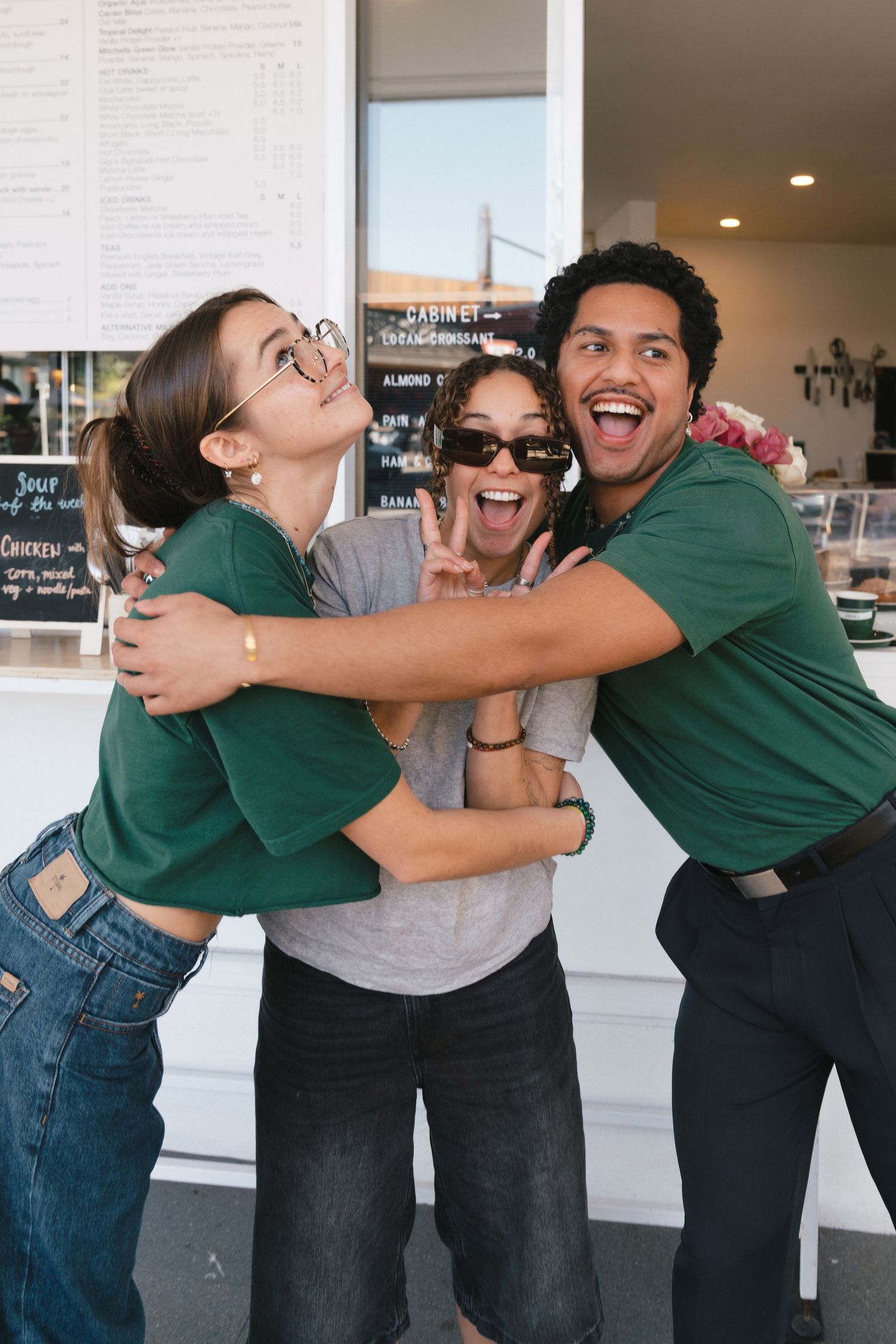Three friends in casual outfits share a joyful group hug and laugh together in front of a cafe counter.