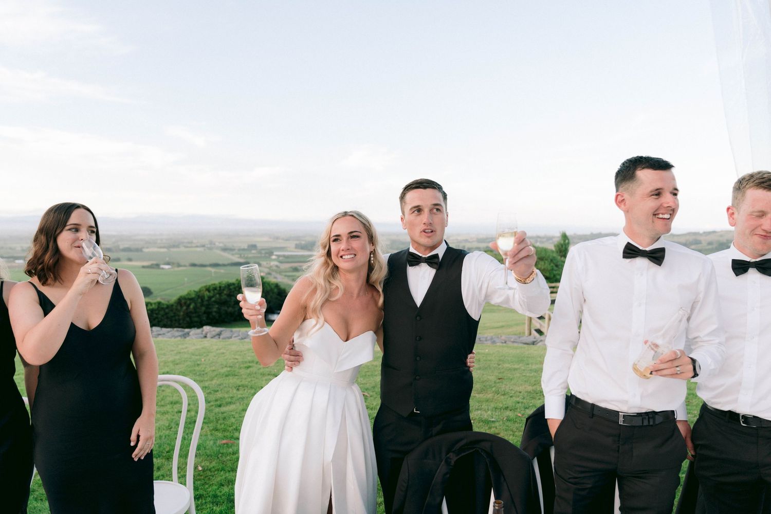 Wedding party in formal black and white attire posing outdoors against a scenic landscape.