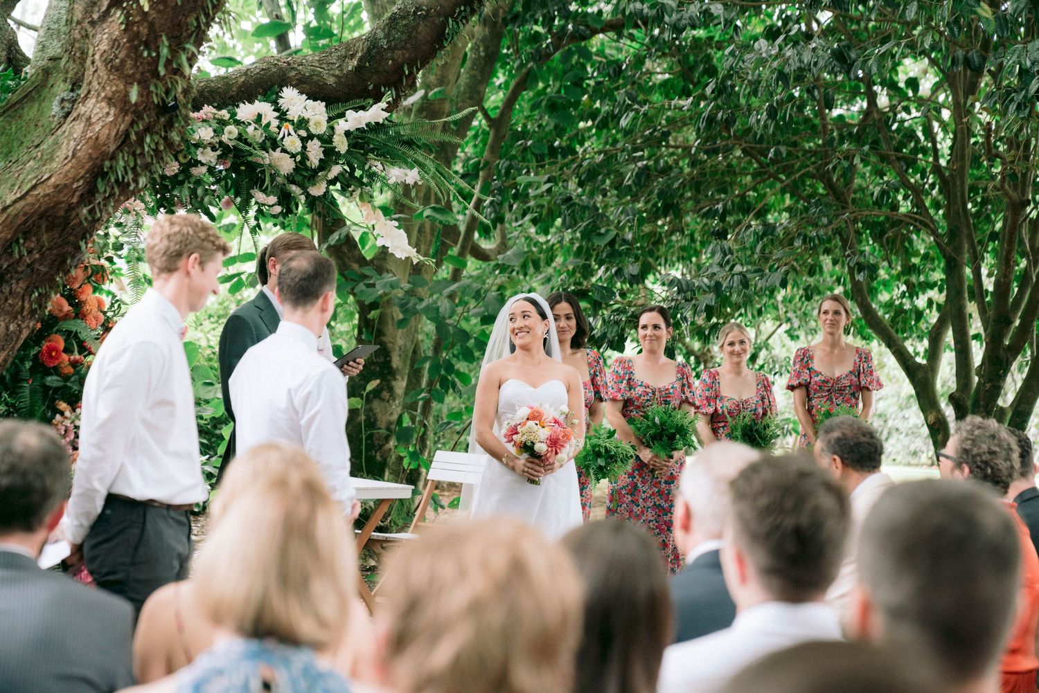 An outdoor wedding ceremony under lush trees with white floral decorations.