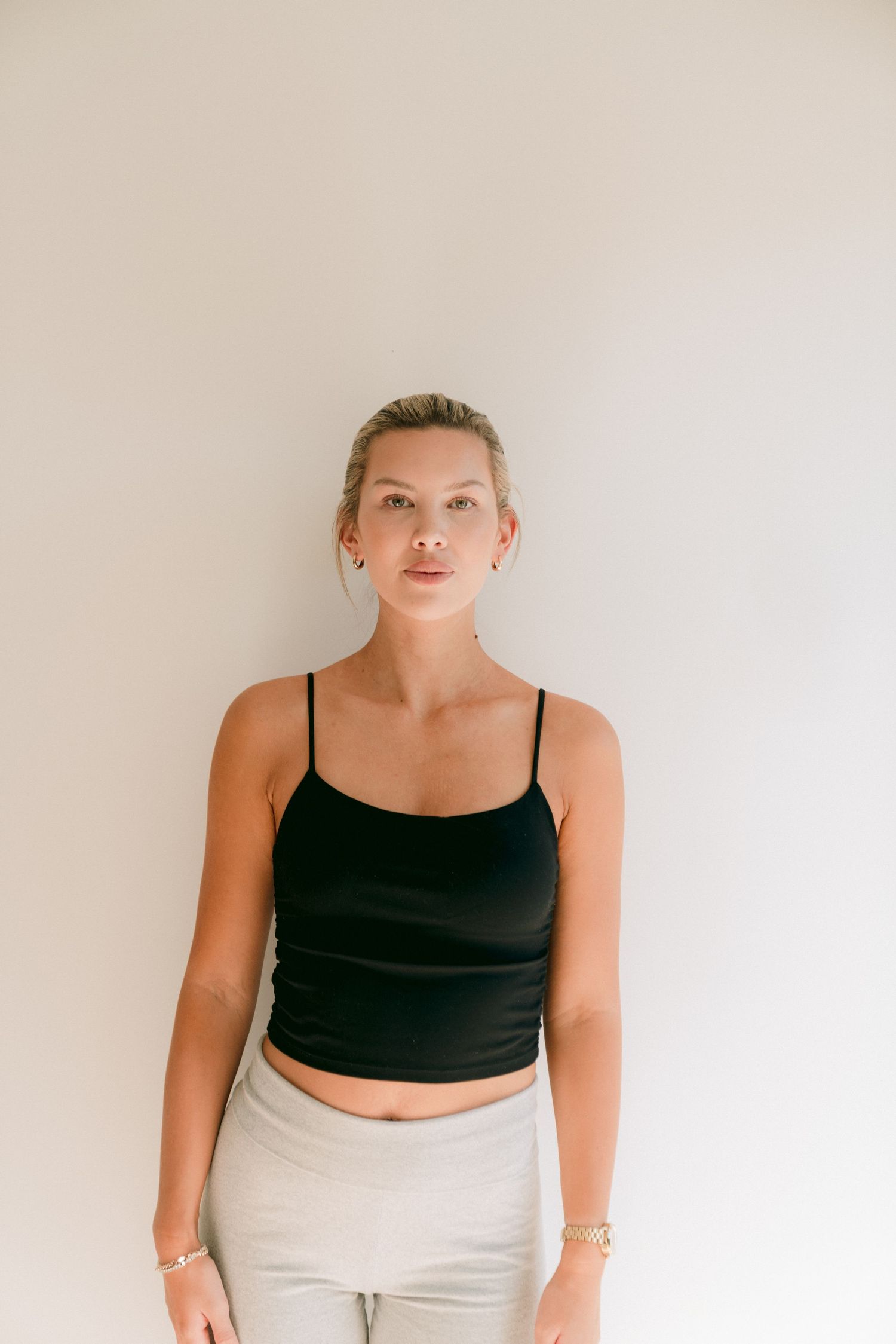 Fashion model wearing black crop top and light pants poses against white wall for casual lifestyle photoshoot.