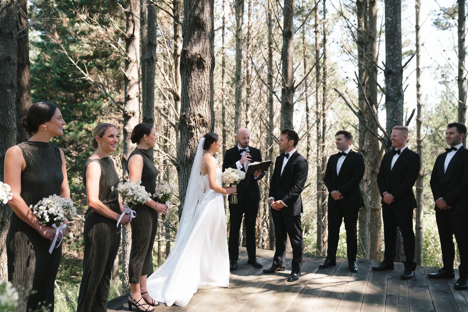 Full wedding party gathered on wooden platform for forest ceremony with pine tree backdrop.