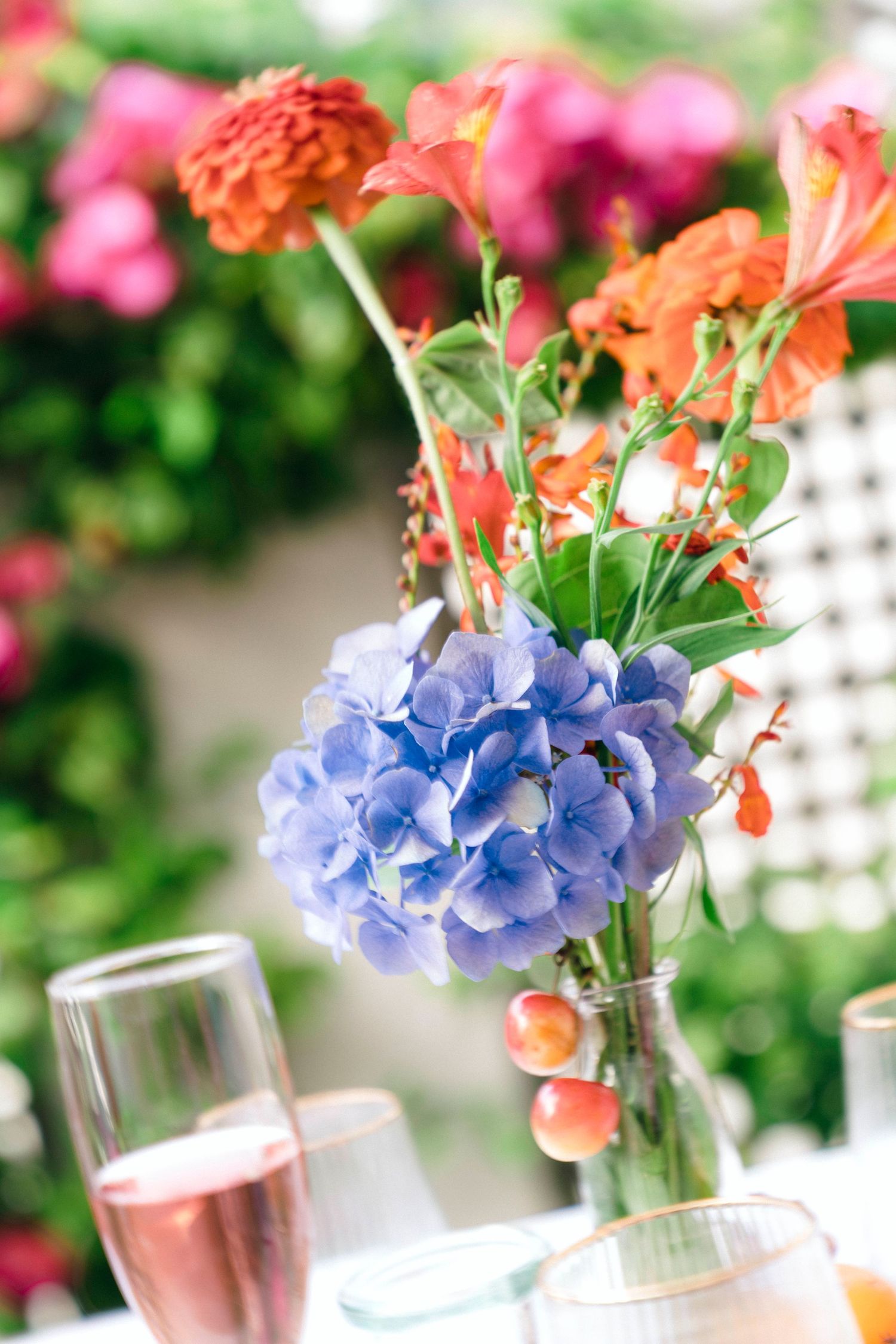 Blue hydrangeas and orange flowers in a vase with champagne glasses on a table.