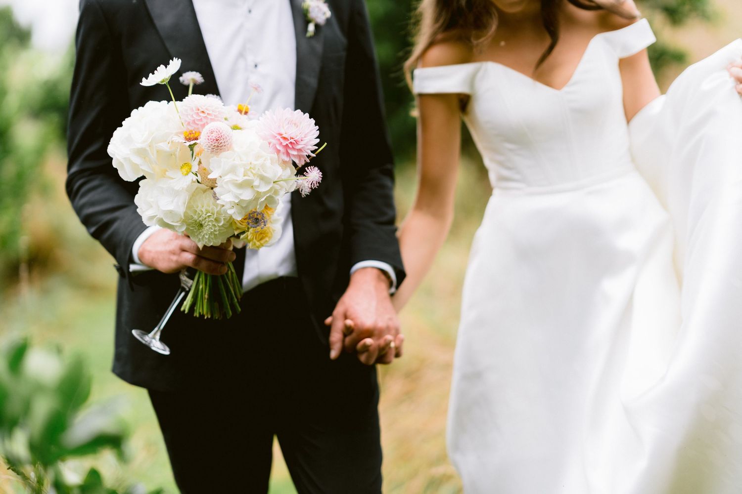 A couple holding hands at a wedding, with the groom in black suit holding a white and pink floral bouquet.