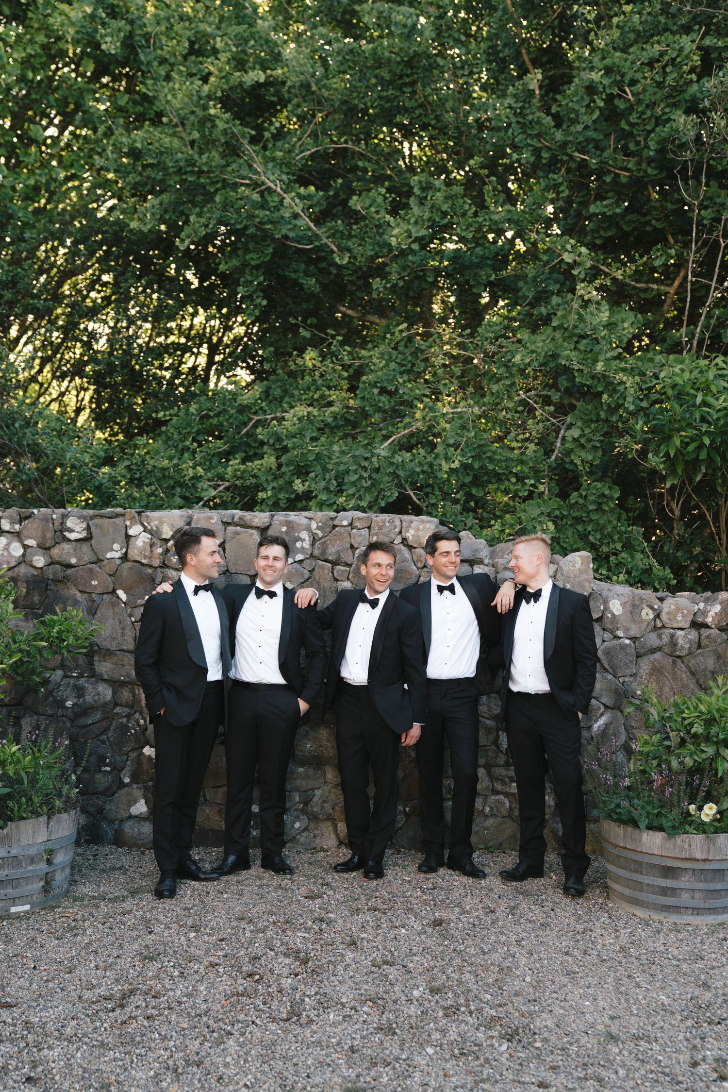 Groomsmen in black tuxedos posing together against rustic stone wall in outdoor garden setting.