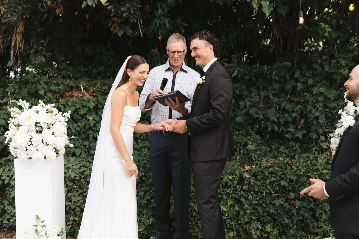 Elegant outdoor wedding ceremony takes place in front of lush greenery wall with white floral arrangements.