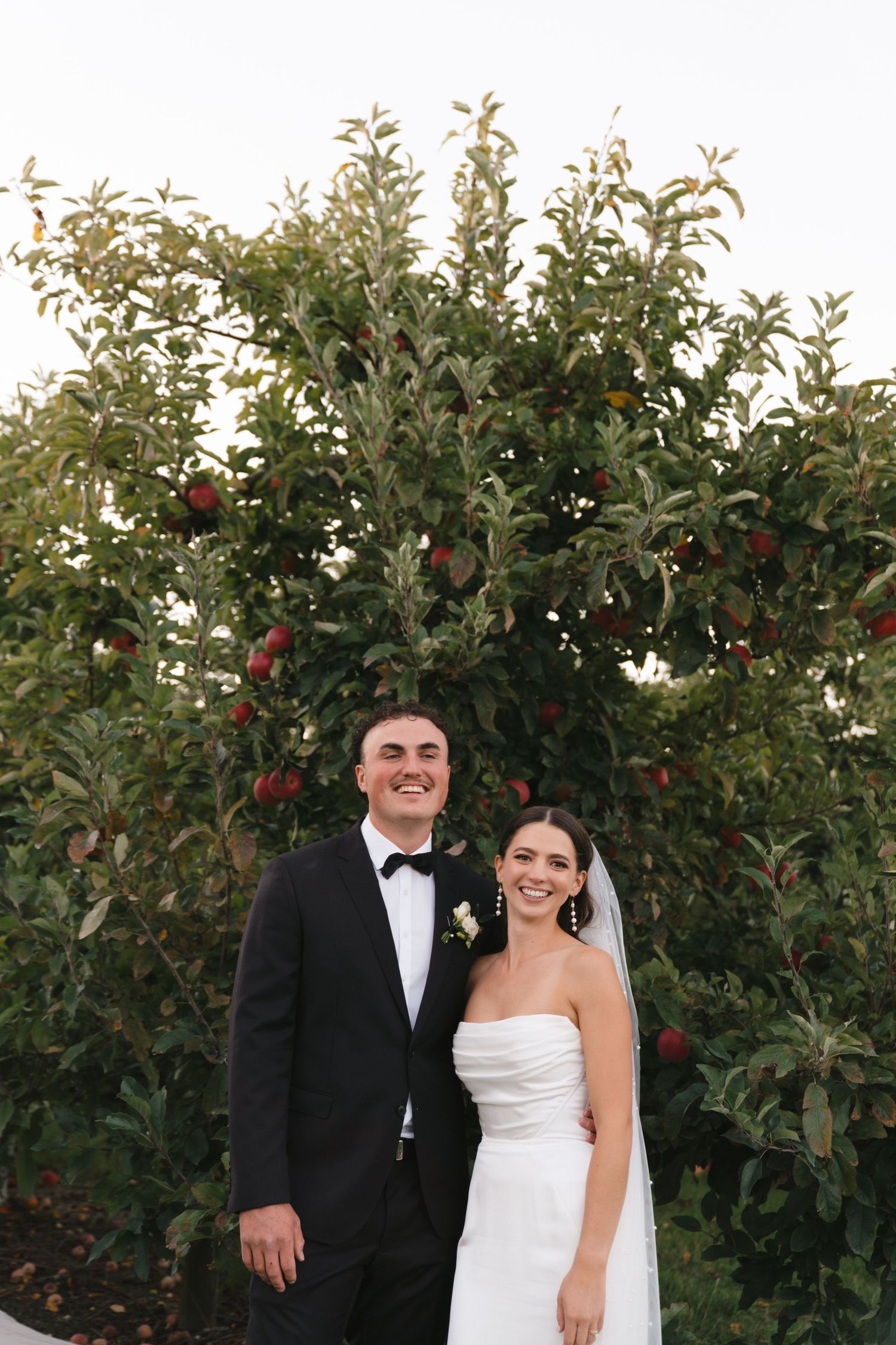 Wedding couple in formal attire standing in front of an apple tree in an orchard.