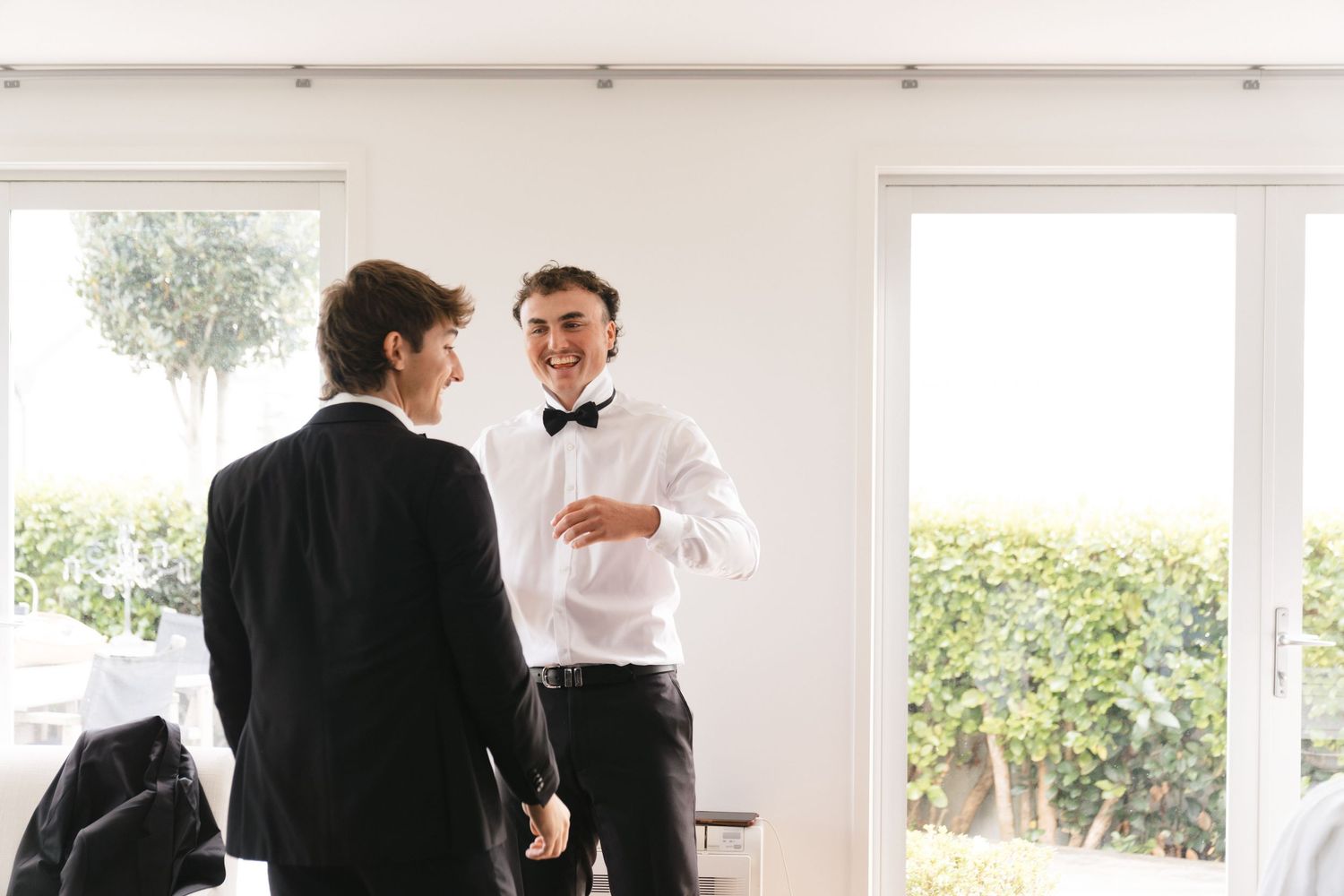 Two people in formal attire having a conversation in a bright indoor space with windows and greenery visible outside.