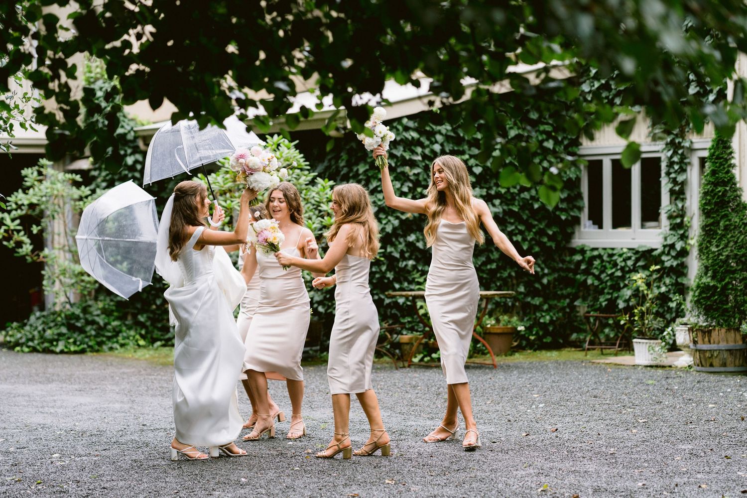 Wedding party in white dresses dancing together on a gravel path surrounded by greenery.