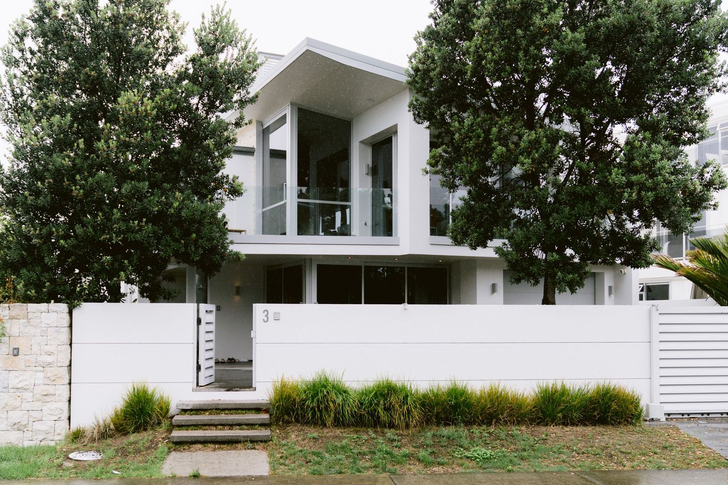 Modern white two-story house with large windows and trees features contemporary architectural design and clean lines.