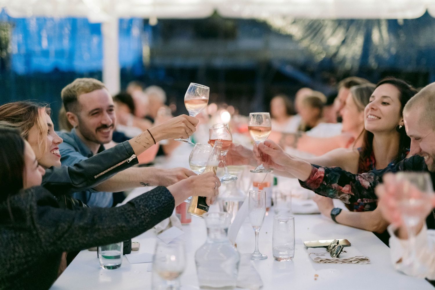 Wedding guests raise their glasses in a celebratory toast at an outdoor reception table.