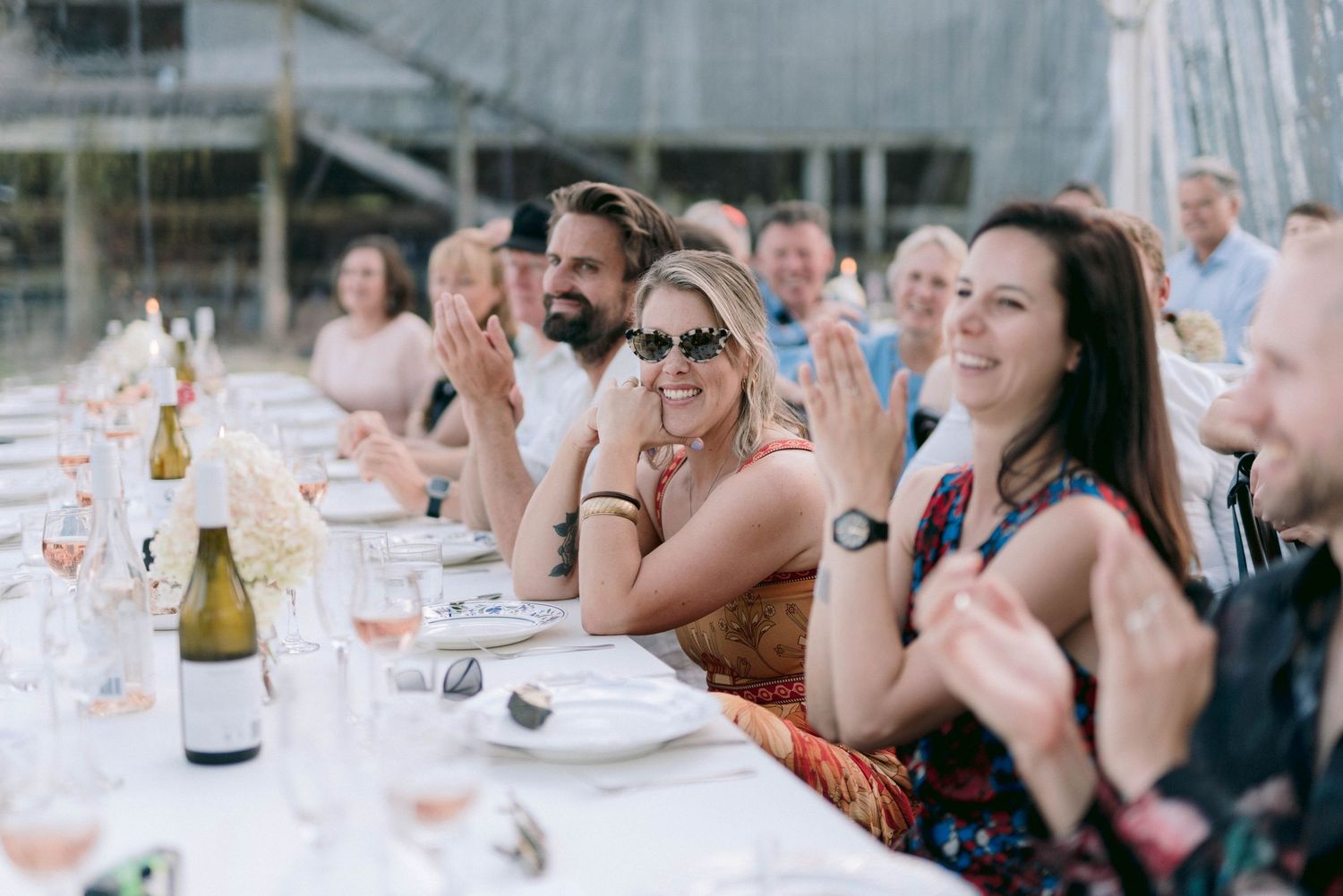Wedding reception guests seated at long tables under a white tent raise their glasses in celebration.