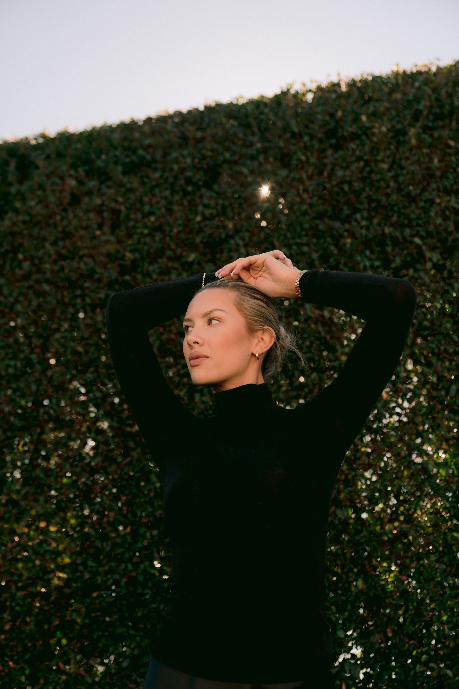 A black-clad figure poses expressively against a dark leafy background at twilight.