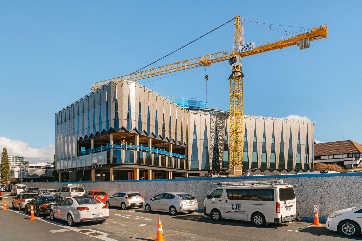 Modern commercial building under construction with a yellow crane and parked cars in the foreground against blue sky.