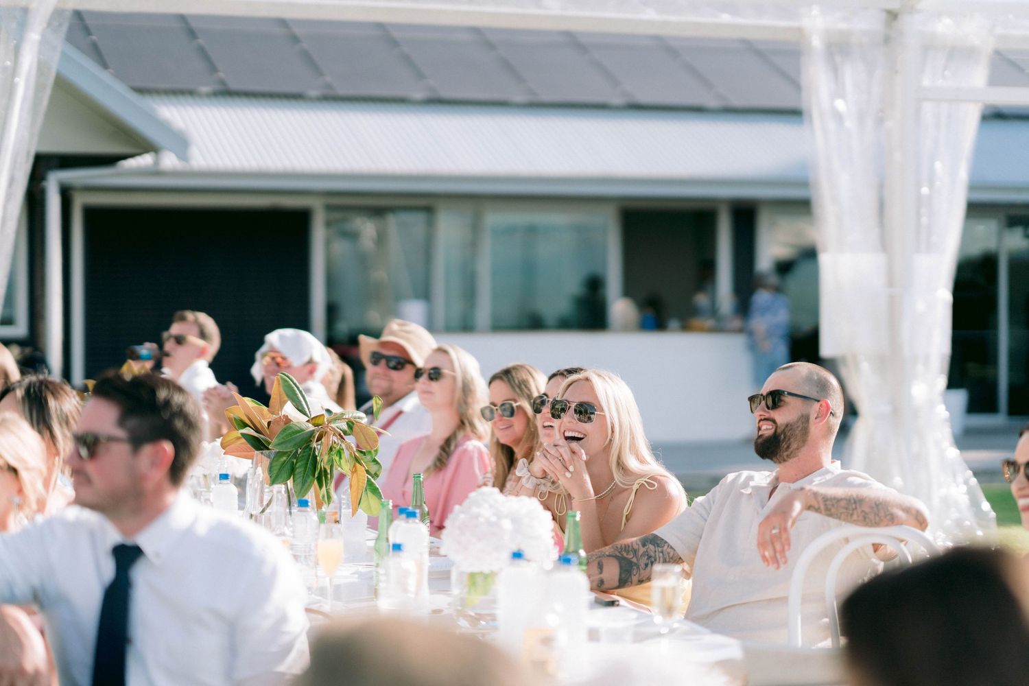 Wedding reception guests seated at outdoor tables under a white tent canopy.