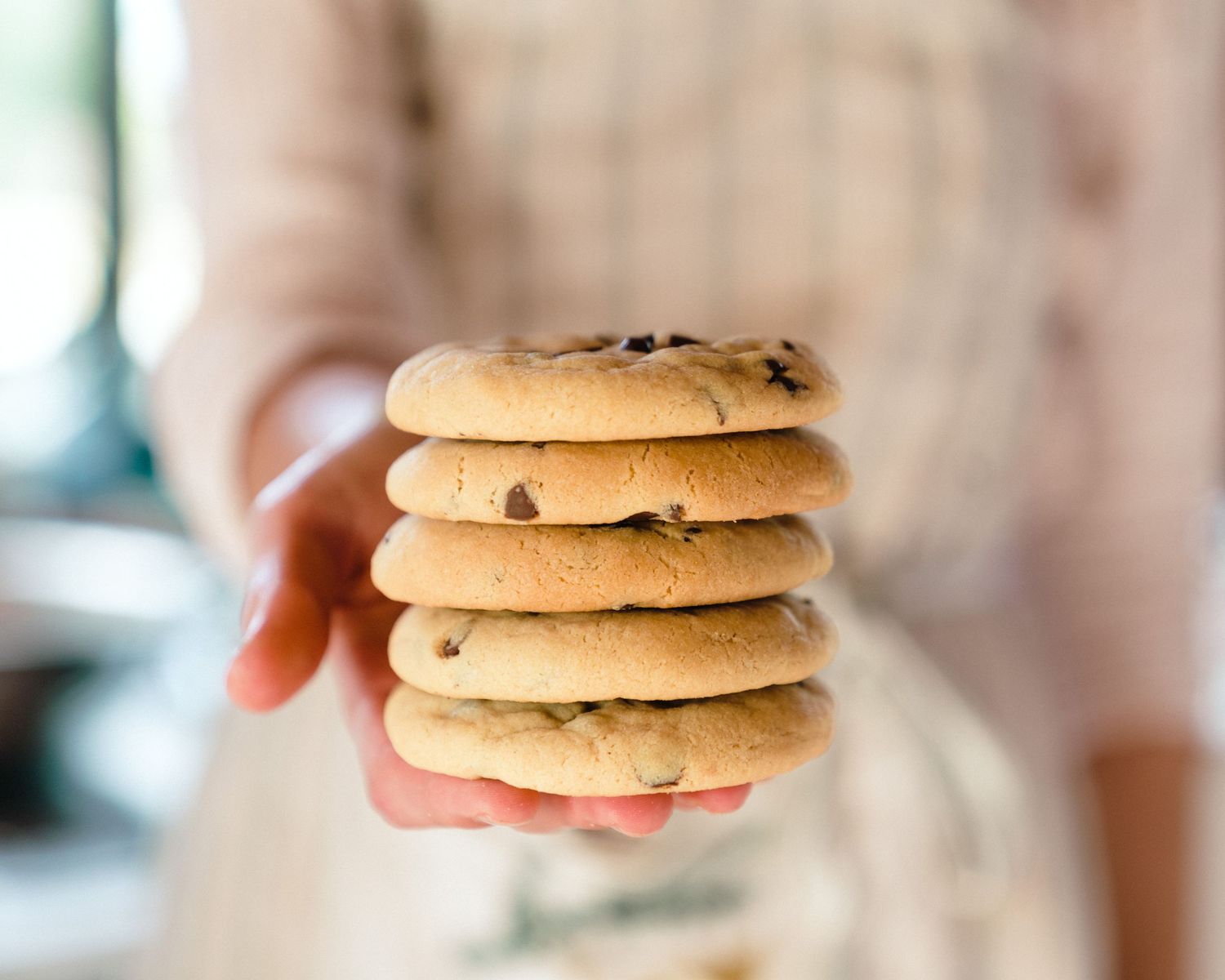 A stack of five chocolate chip cookies being held up against a blurred background.