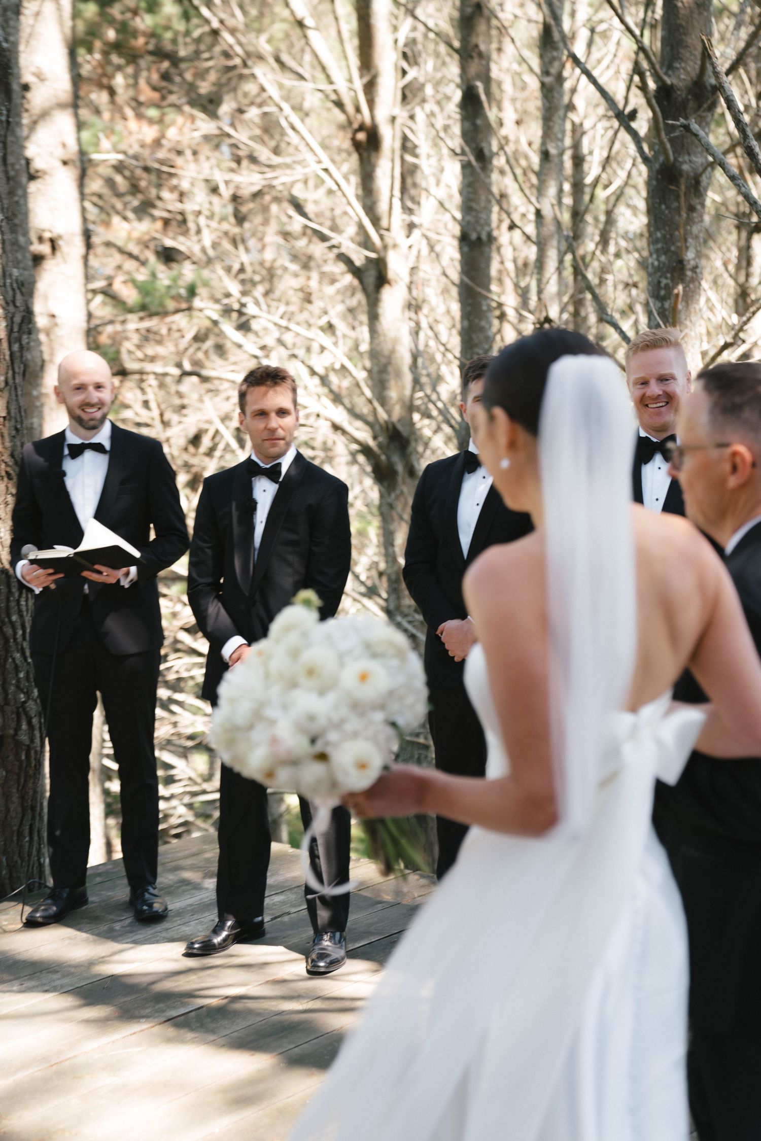 Outdoor forest wedding ceremony with attendants in formal black tie attire.