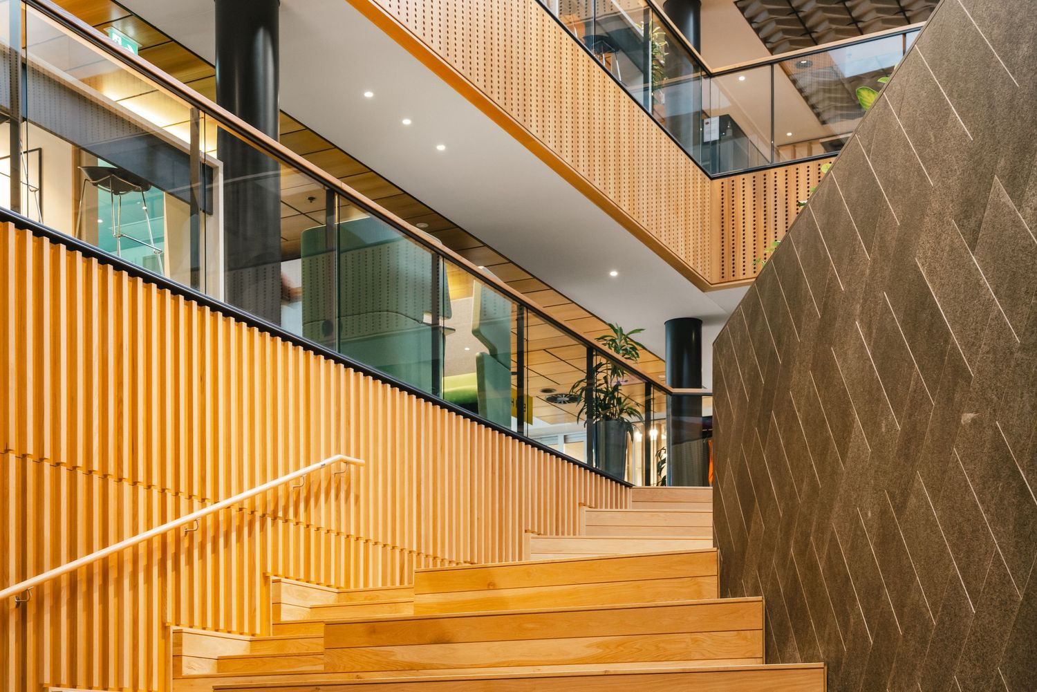 Modern office interior features wooden stairs with glass railings and vertical timber slat walls illuminated by recessed lighting.