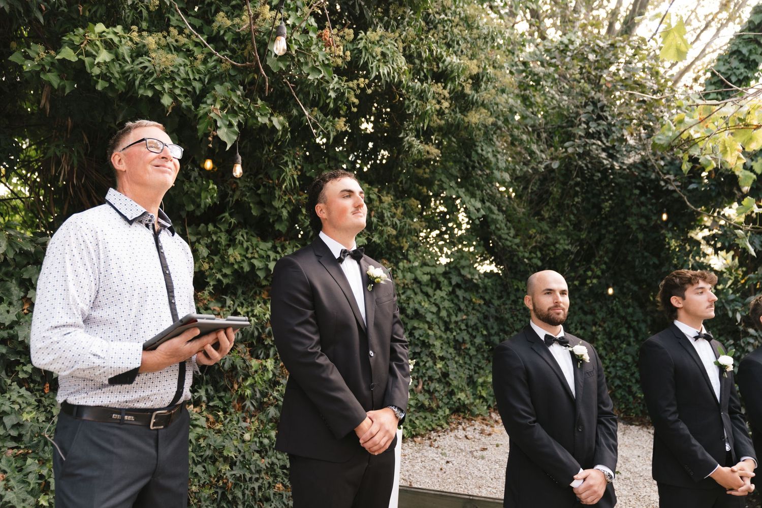 Four people in formal attire stand in line at an outdoor wedding ceremony with greenery in the background.