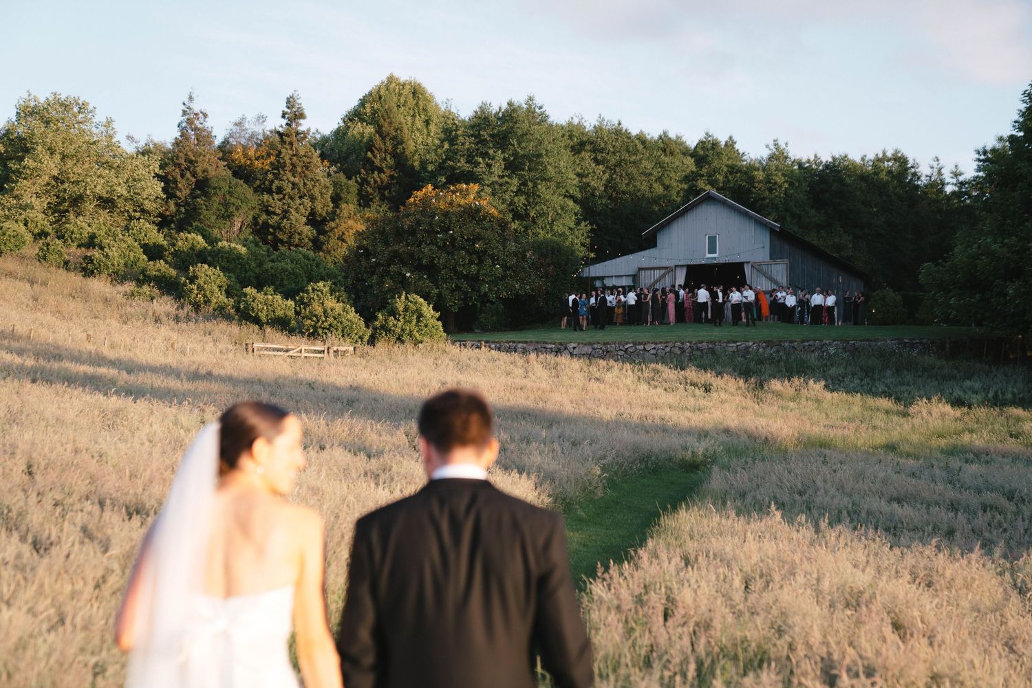 A couple walking through a grassy field toward a rustic white farmhouse on a hill at sunset.