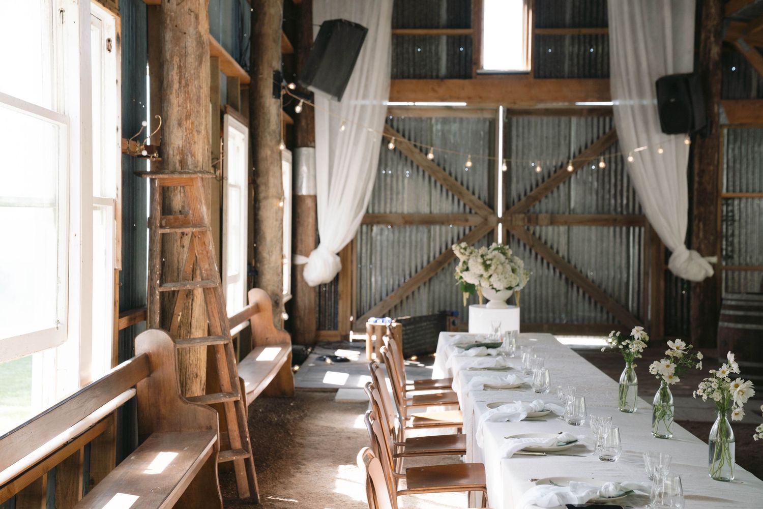 A rustic barn wedding reception setup with long wooden tables, white linens, and string lights in an elegant farmhouse venue.