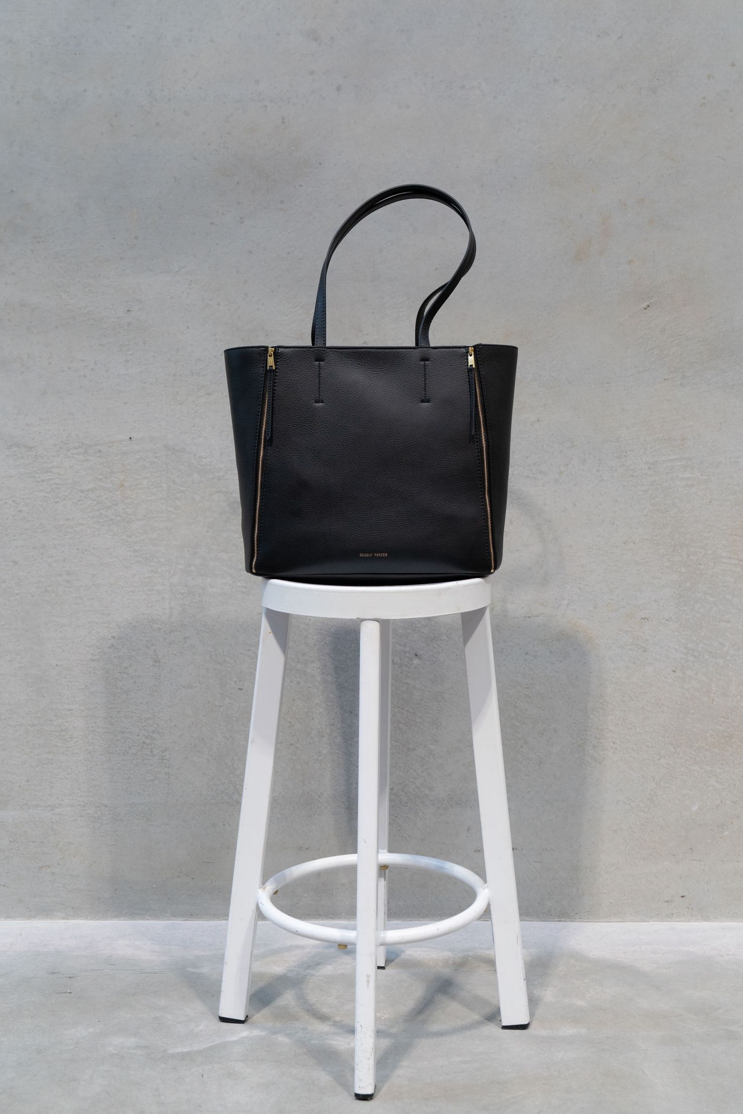 A black leather tote bag with gold accents sits atop a minimalist white metal bar stool against a gray wall.