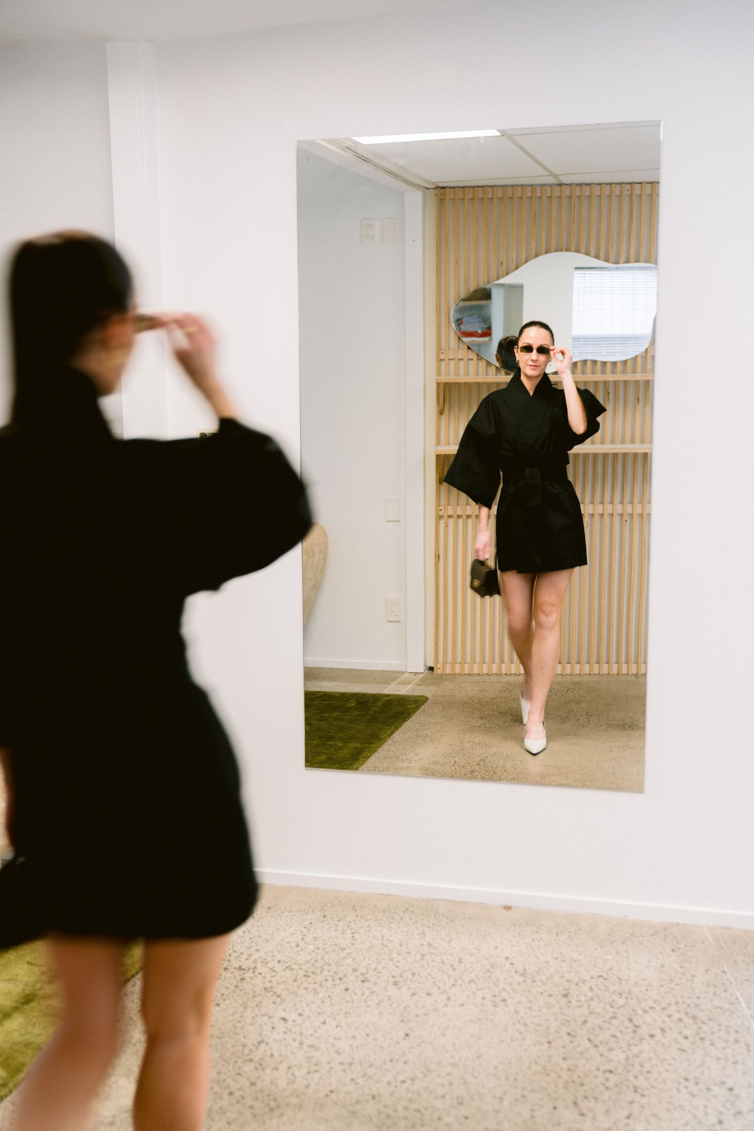 Someone wearing a black mini dress and white shoes poses in front of a mirror while taking a photo.