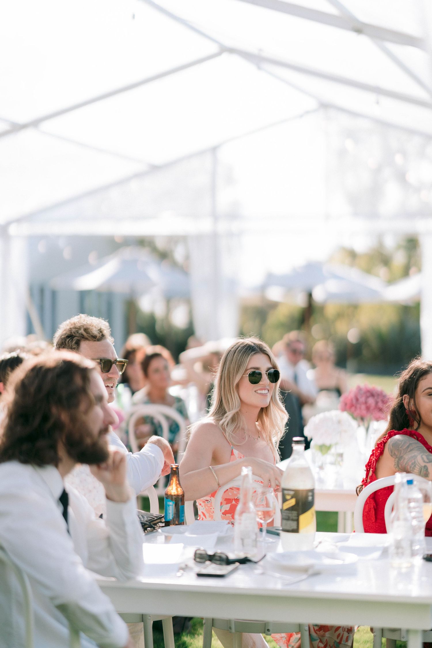 Wedding guests enjoying an outdoor reception under a white tent with tables and floral centerpieces.