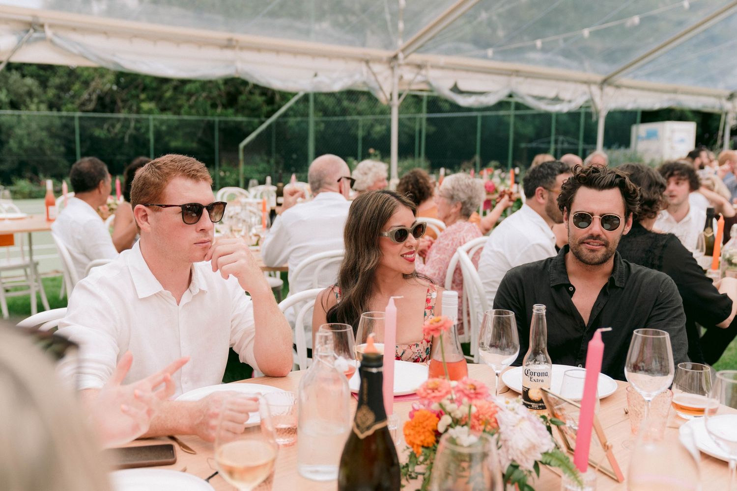 Wedding guests enjoying drinks and conversation at an outdoor reception under a clear tent.