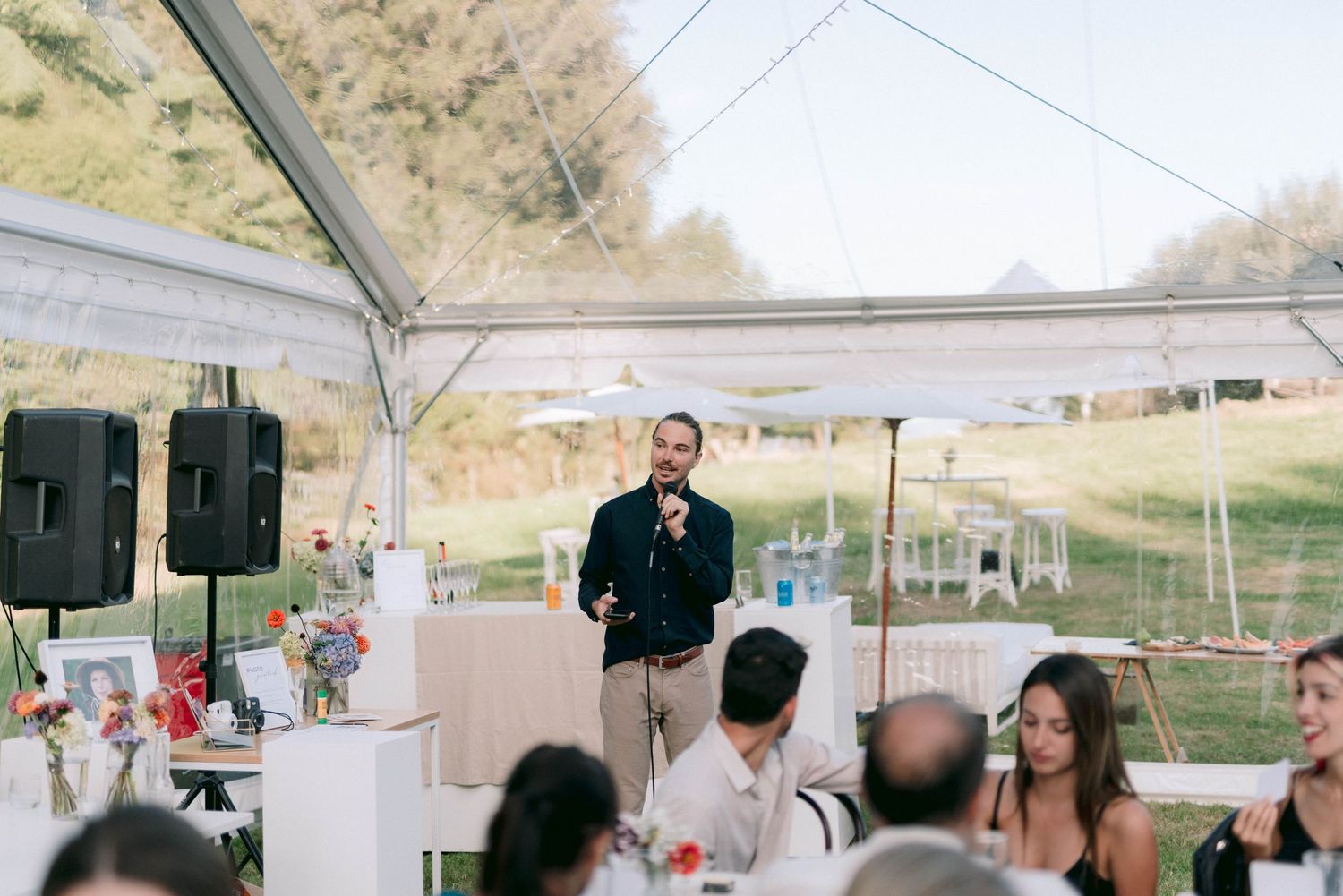 Wedding reception speaker addressing guests under a white tent with speakers and decorations.