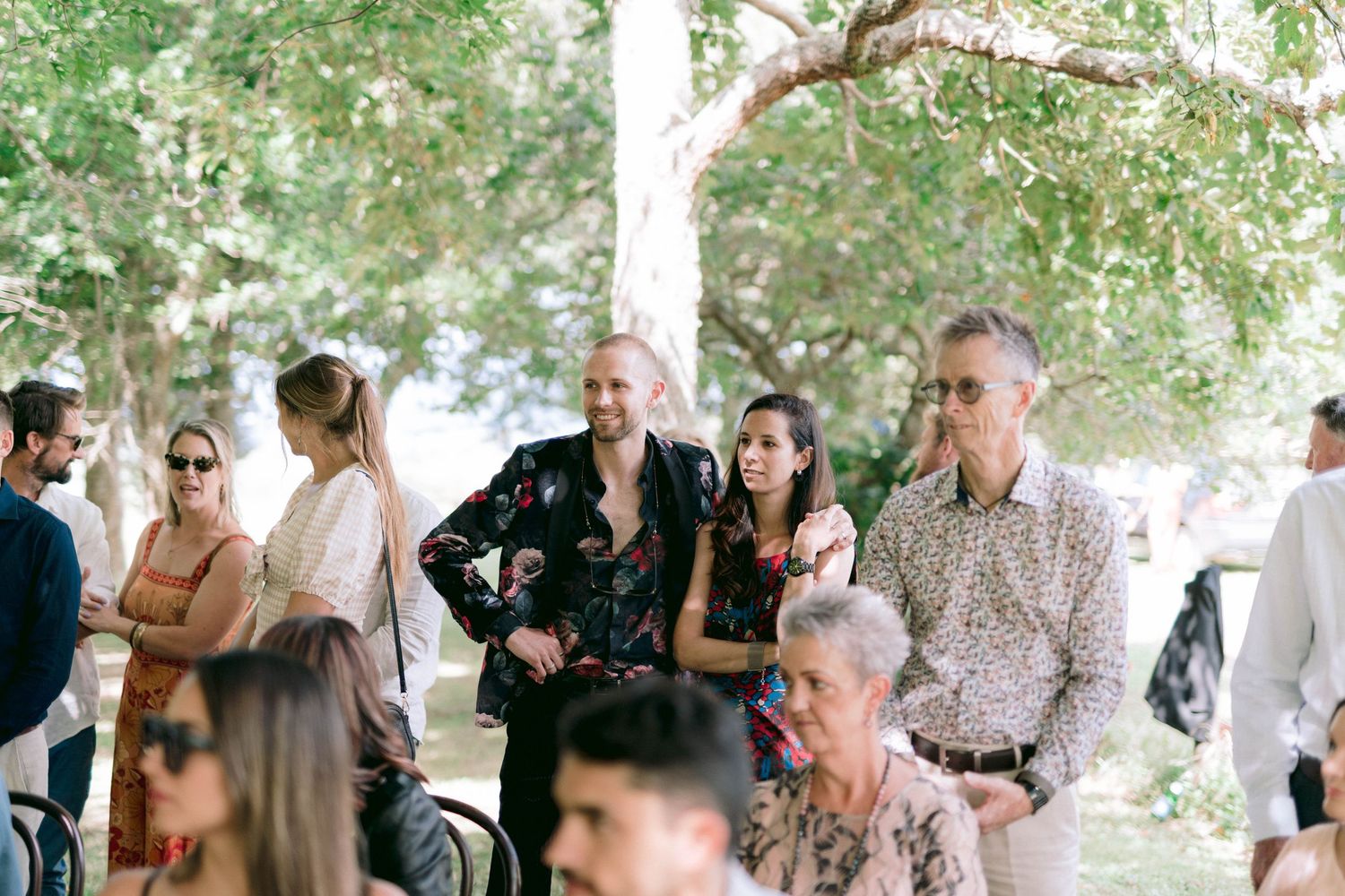 Outdoor gathering of wedding guests under trees with natural summer lighting.