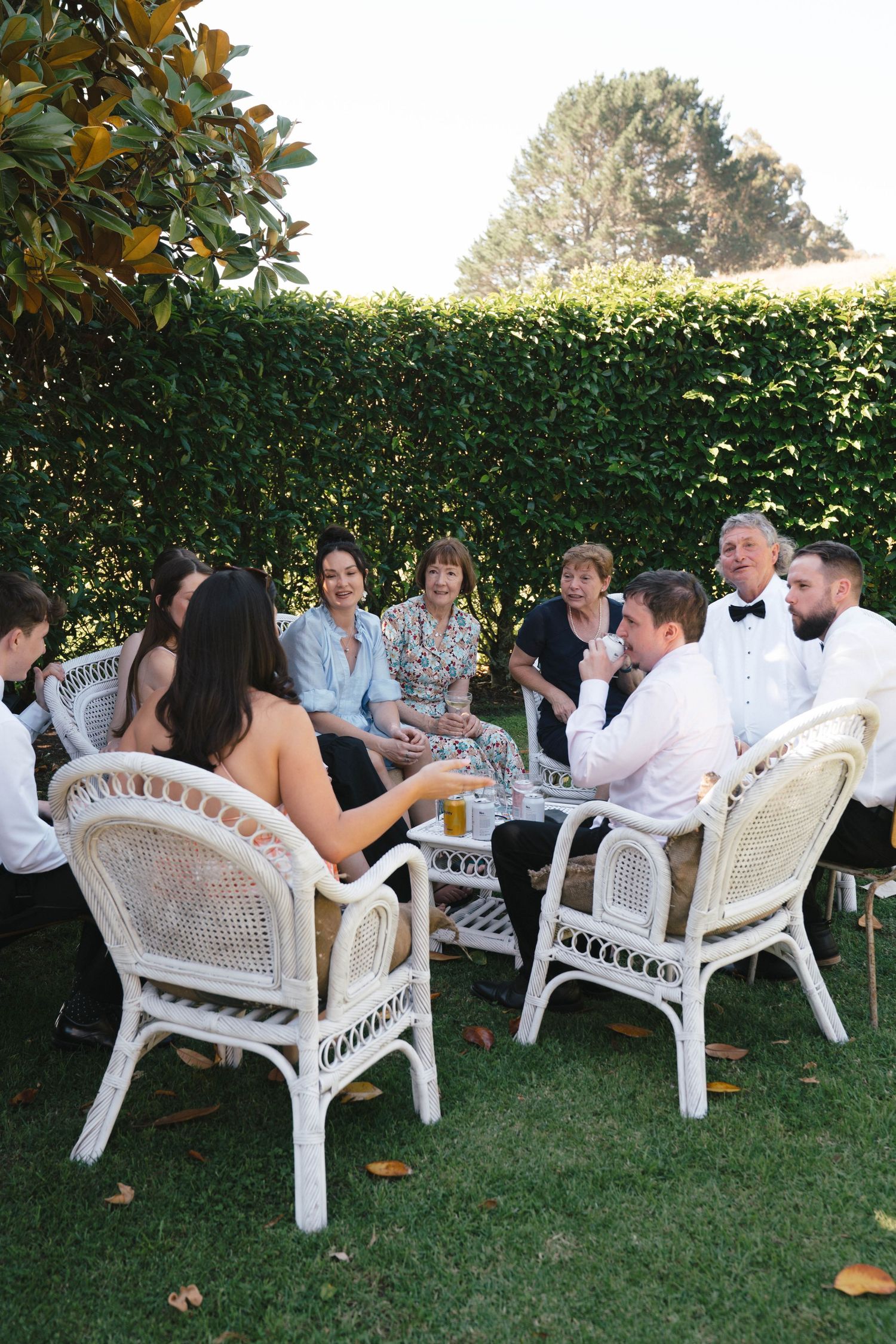 A group sits together at white wicker chairs around a table for an outdoor garden party surrounded by manicured hedges.