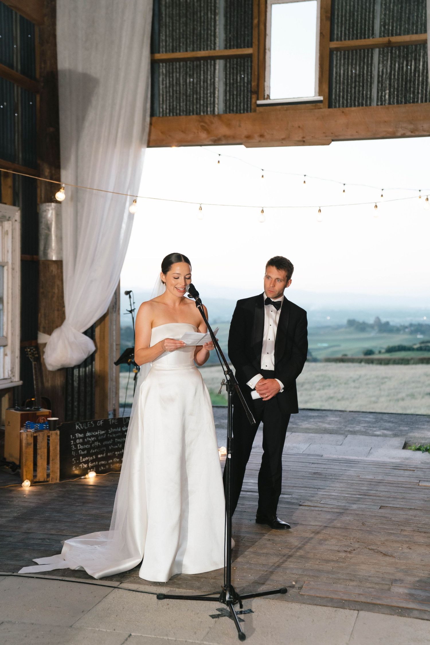Bride giving wedding speech at microphone while groom looks on in rustic barn venue.
