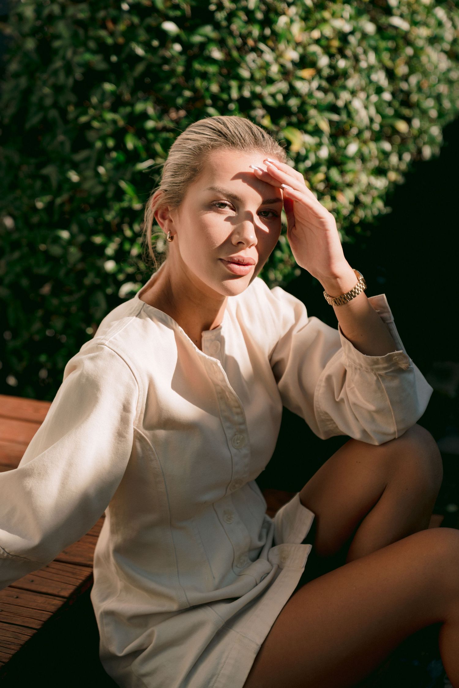 Person in white button-down shirt sitting outdoors in sunlight with hand on forehead in relaxed pose.