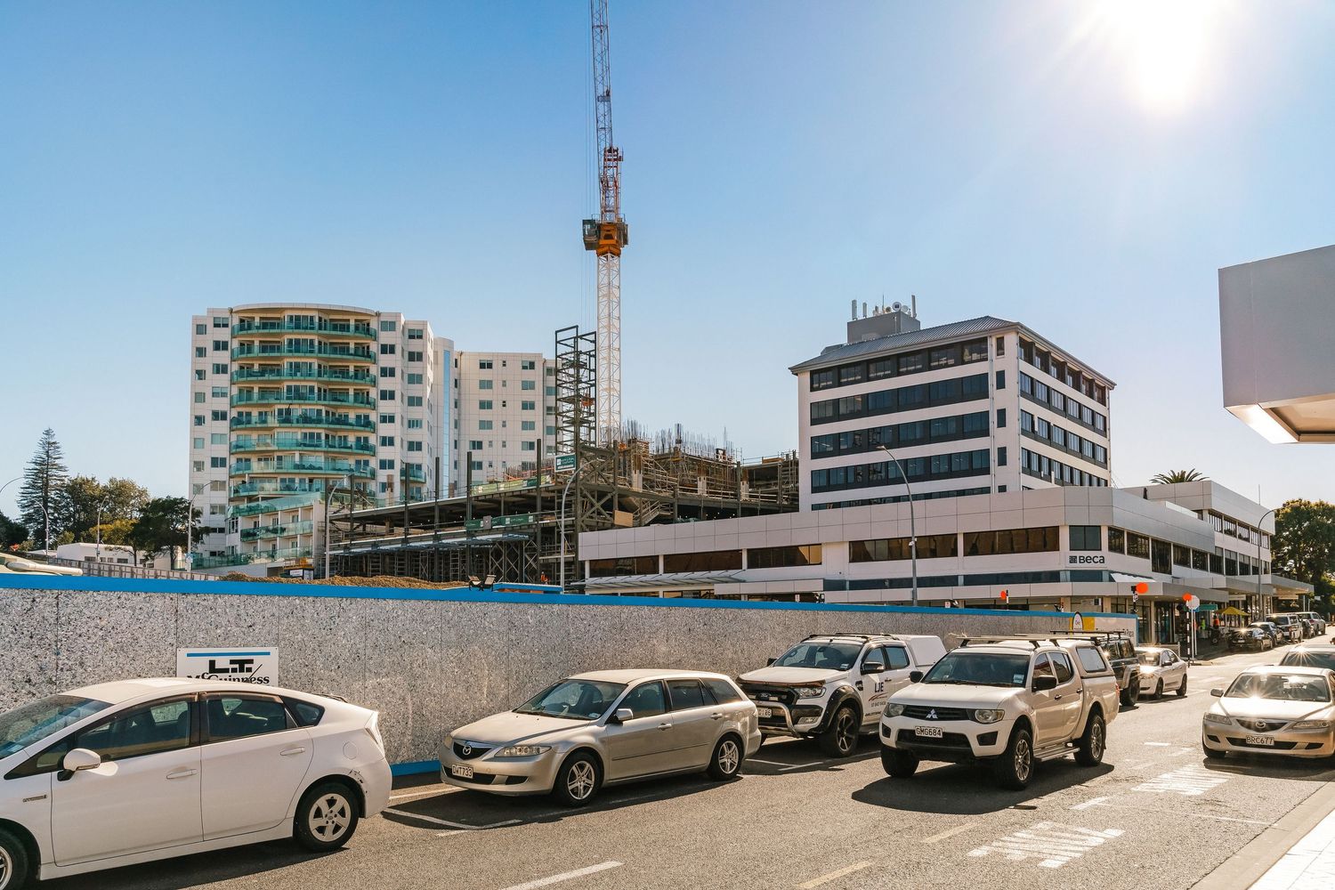 Row of parked cars in front of coastal apartment buildings and communications tower under bright sunlight.