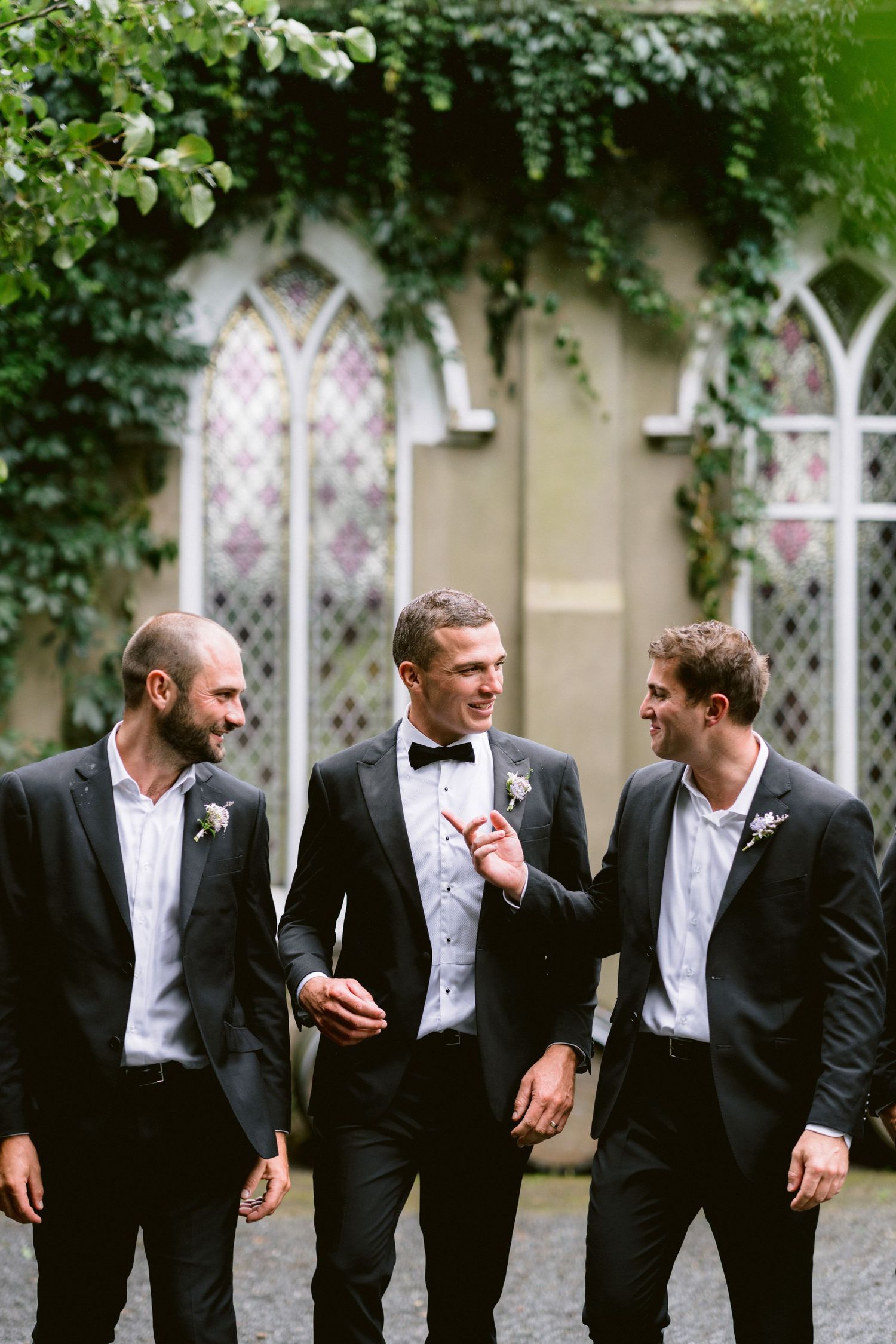 Three groomsmen in black suits sharing a laugh against an ivy-covered wall.