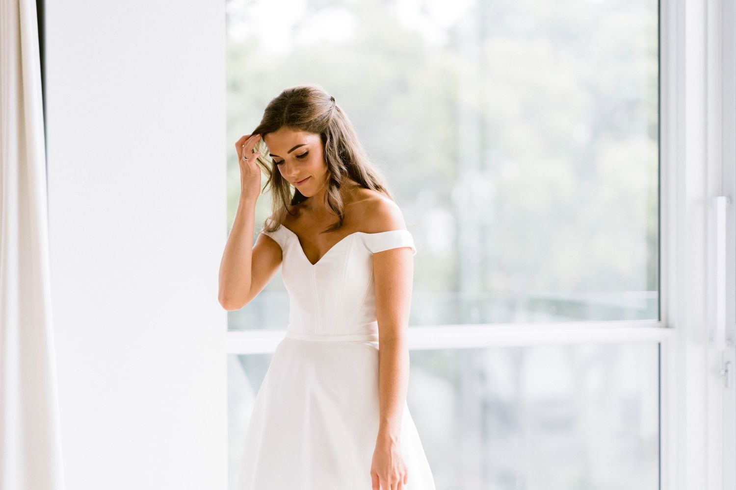 Bride in elegant off-shoulder white wedding dress stands by bright window in natural light.