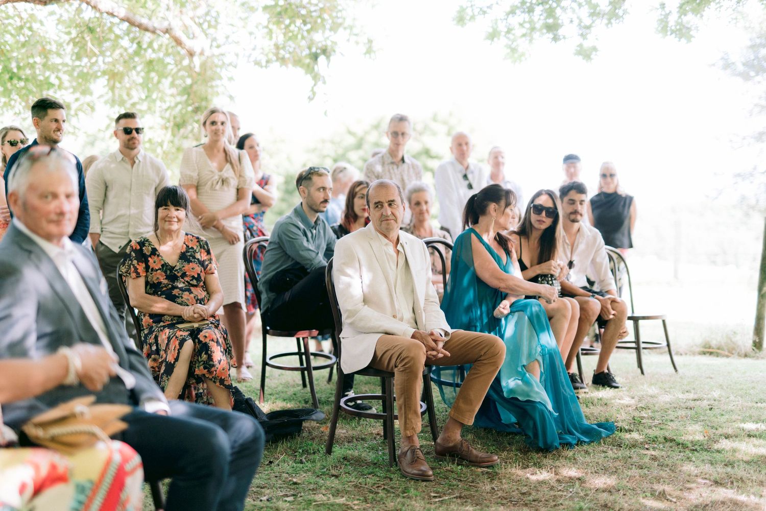 Wedding guests seated on chairs watch an outdoor ceremony under trees.