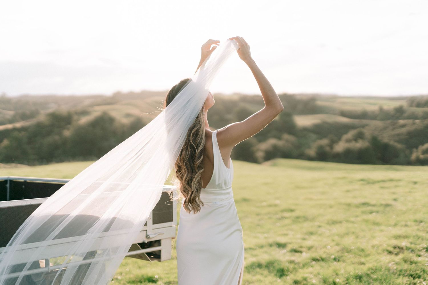 Bride in white dress holding her flowing veil against scenic rolling hills backdrop.