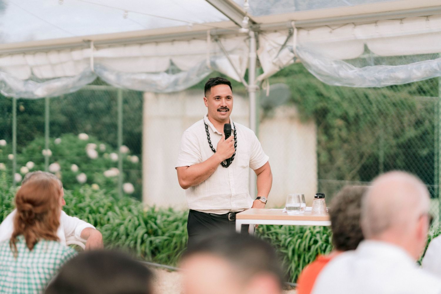 Speaker addressing guests at outdoor wedding reception under white tent.