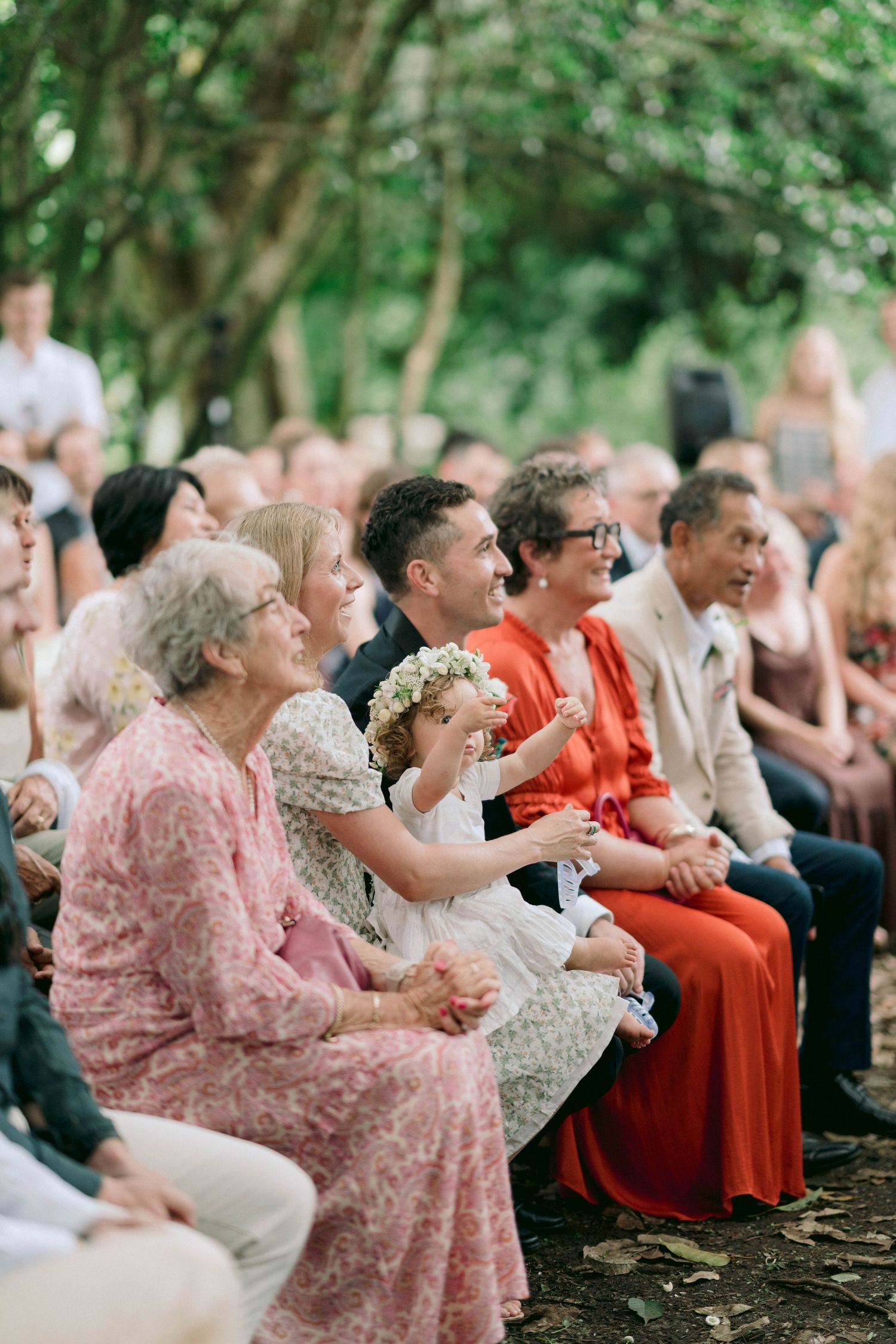 Wedding guests seated on wooden benches watch attentively during an outdoor ceremony.
