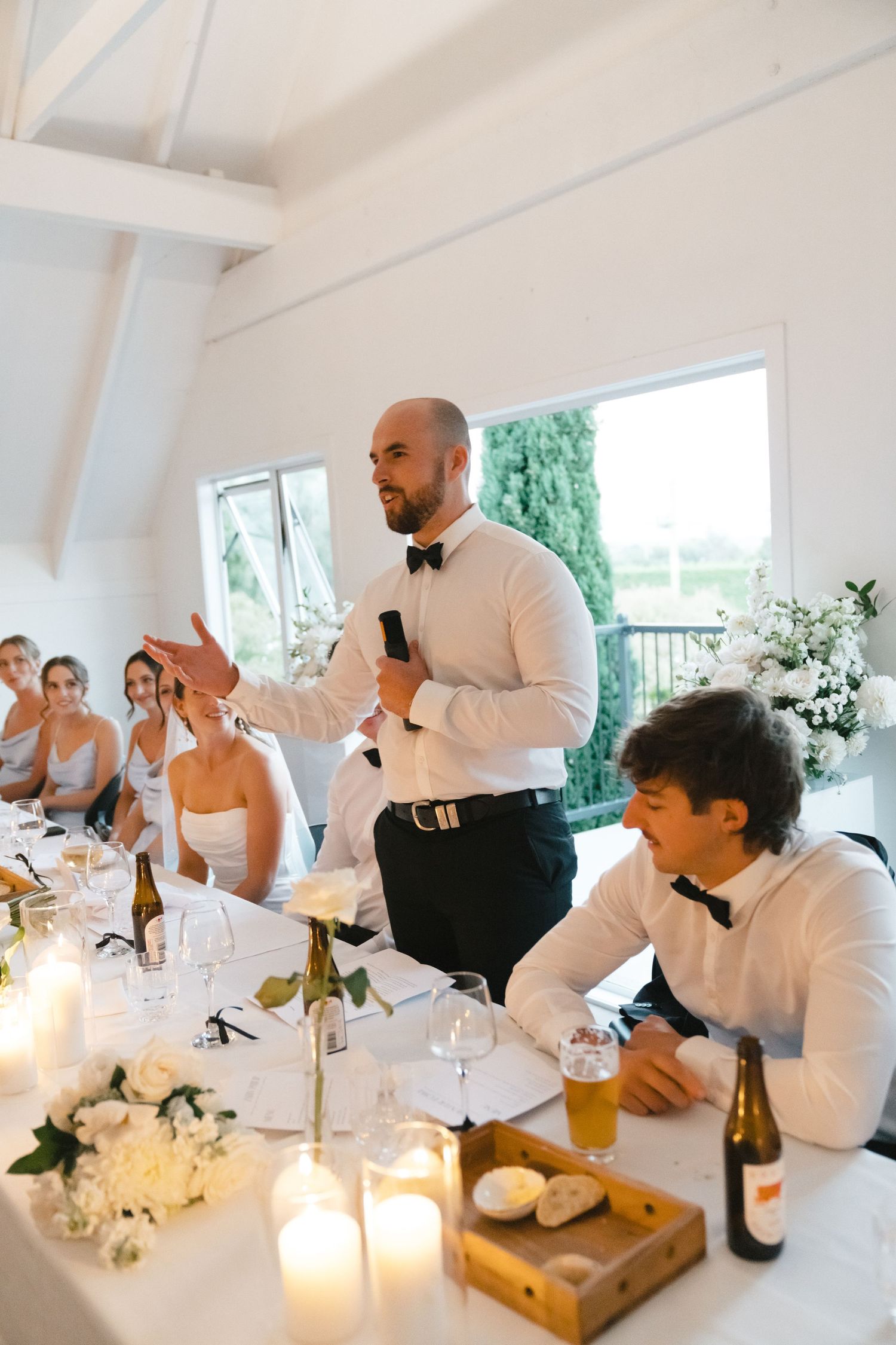 Wedding reception tables set with white decor as guests in formal attire gather around to celebrate.
