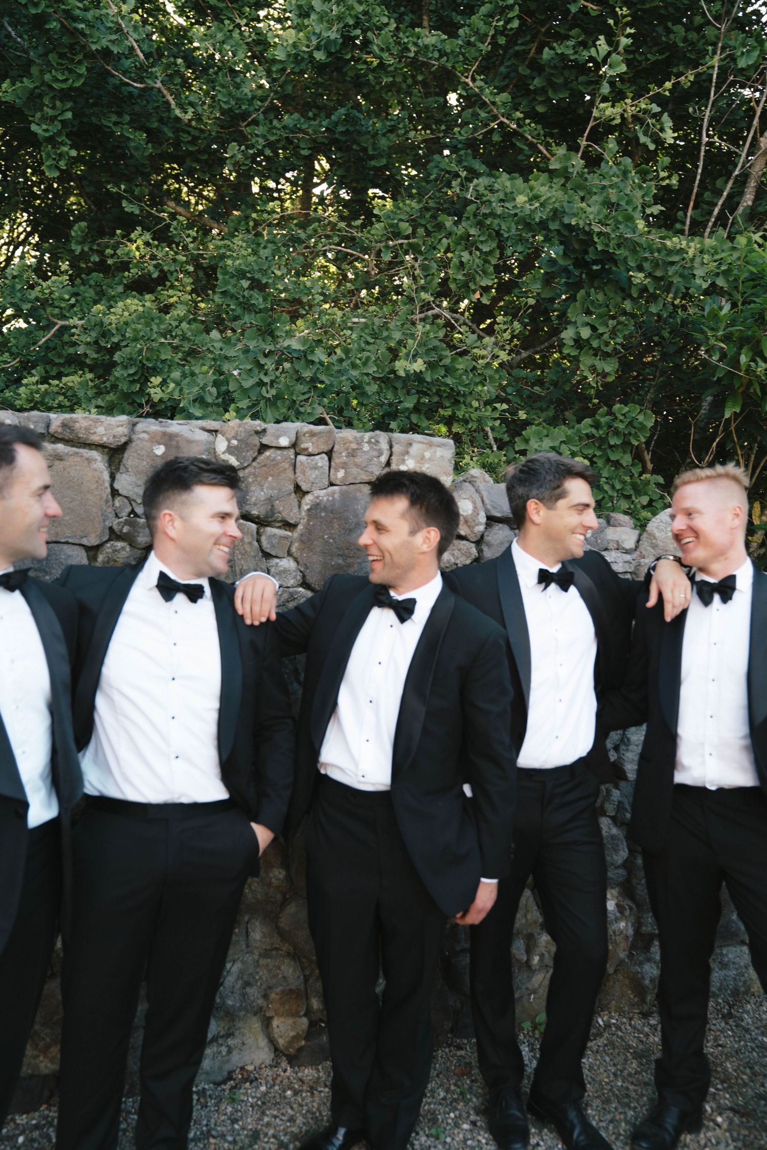 Groomsmen in black tuxedos and bow ties laughing together against a stone wall.