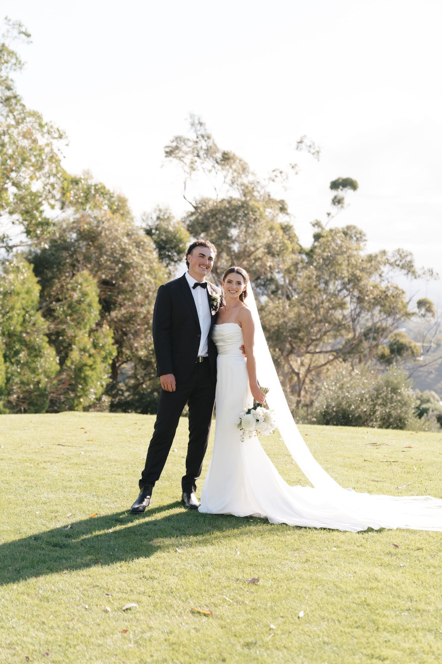 A wedding couple poses together on a grassy hilltop with trees in the background during golden hour.