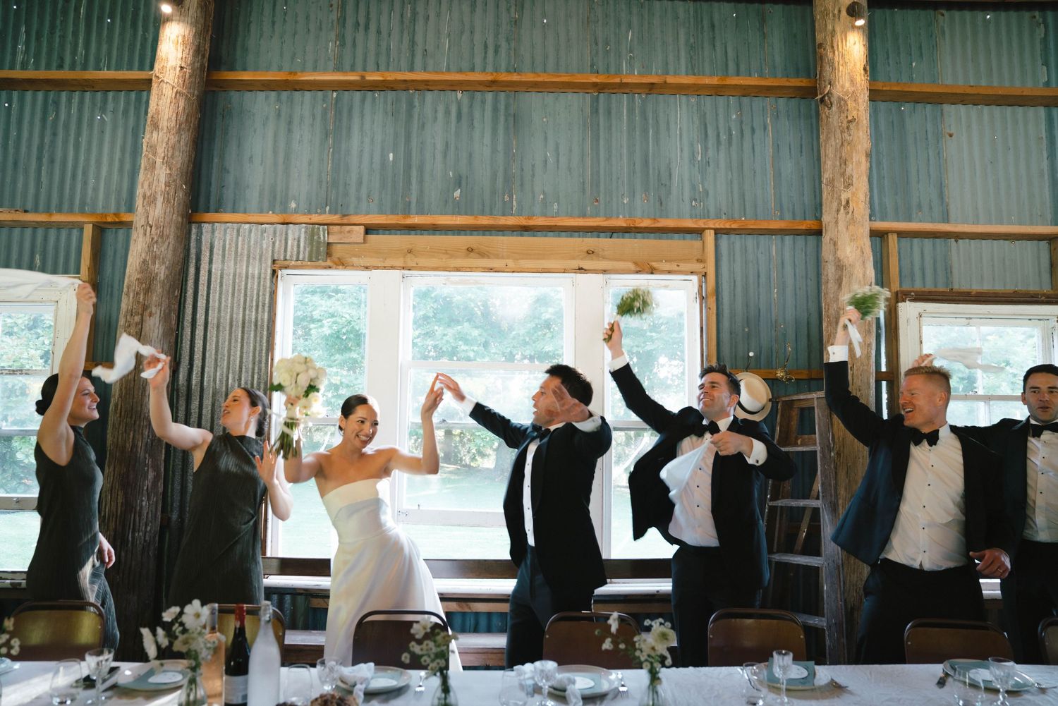 Wedding guests raise their arms in celebration during a joyous reception inside a rustic wooden barn venue.