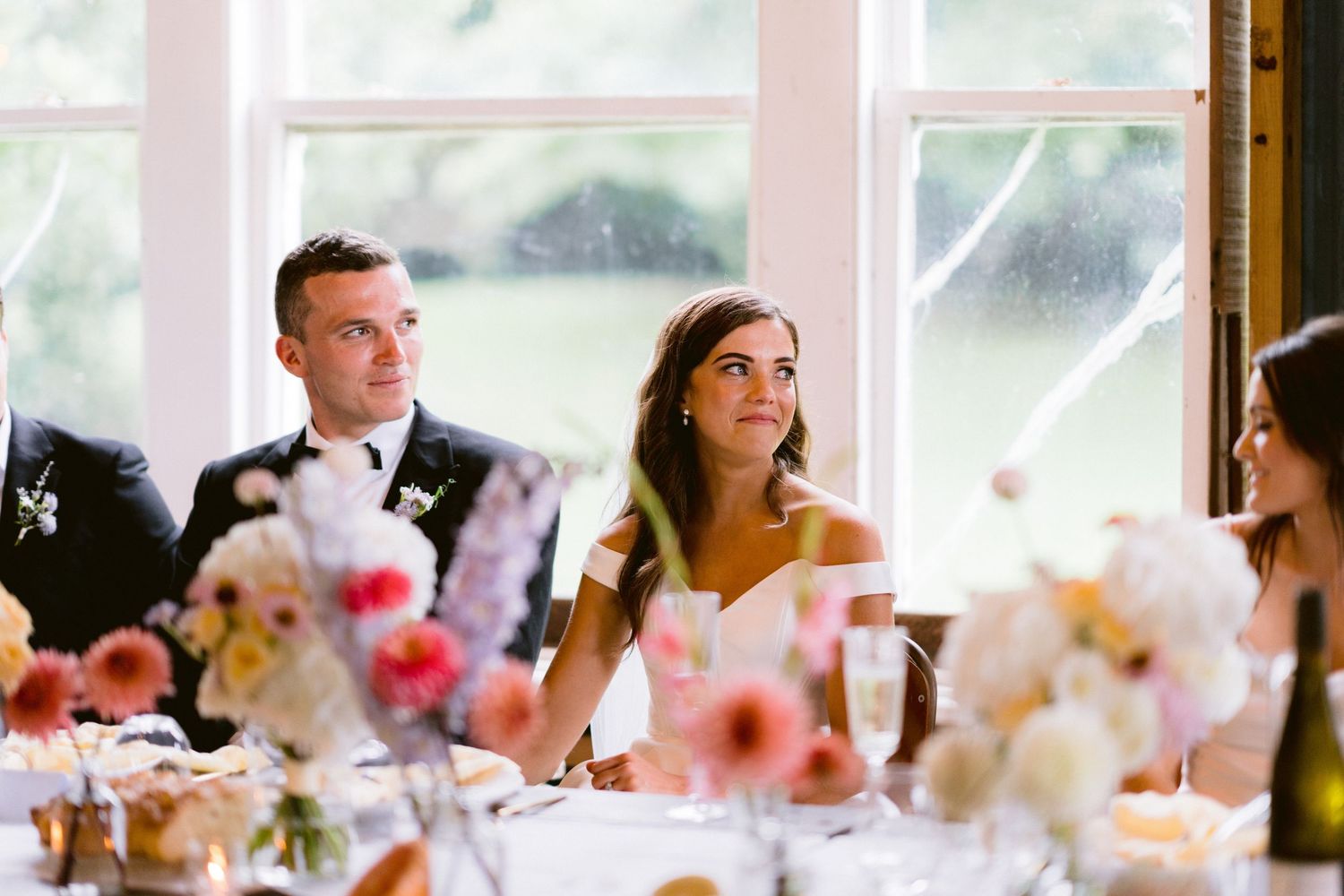 Wedding guests seated at long tables with pink and purple floral centerpieces during an indoor reception.