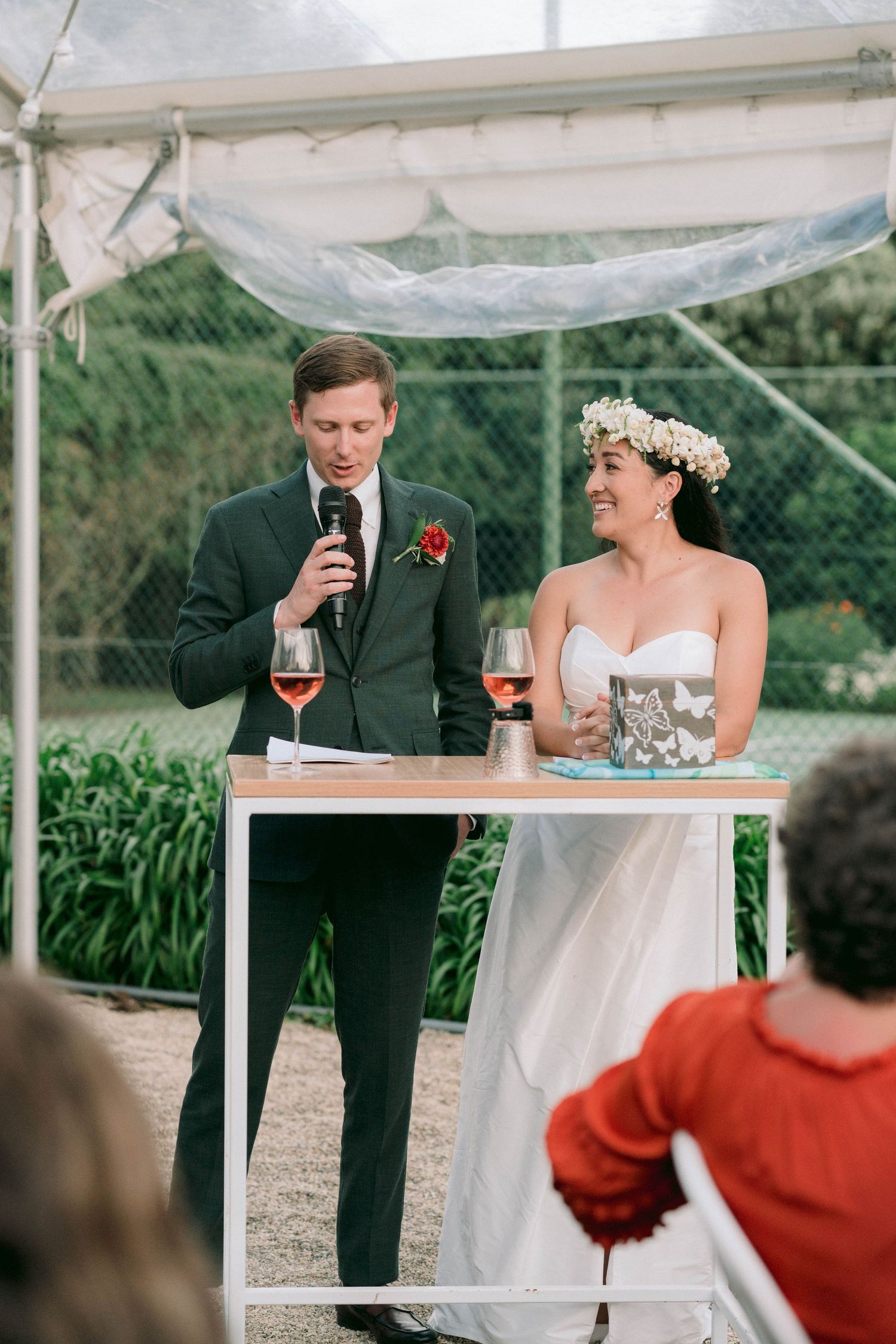 A wedding celebration under a tent with the couple sharing drinks and speeches at a podium.
