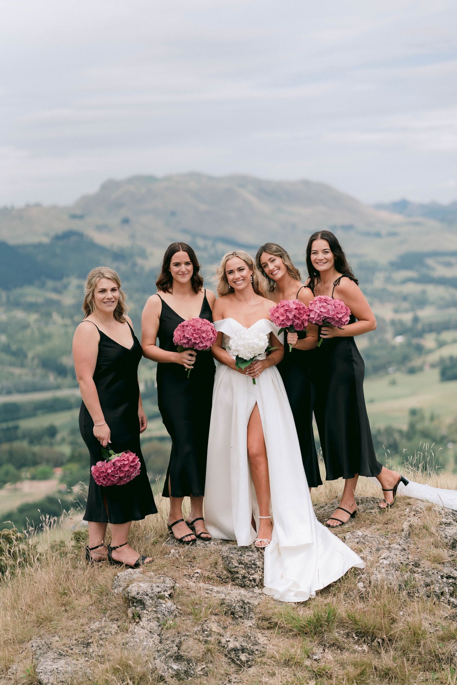 Bridal party with pink bouquets posing on mountaintop with scenic valley view.