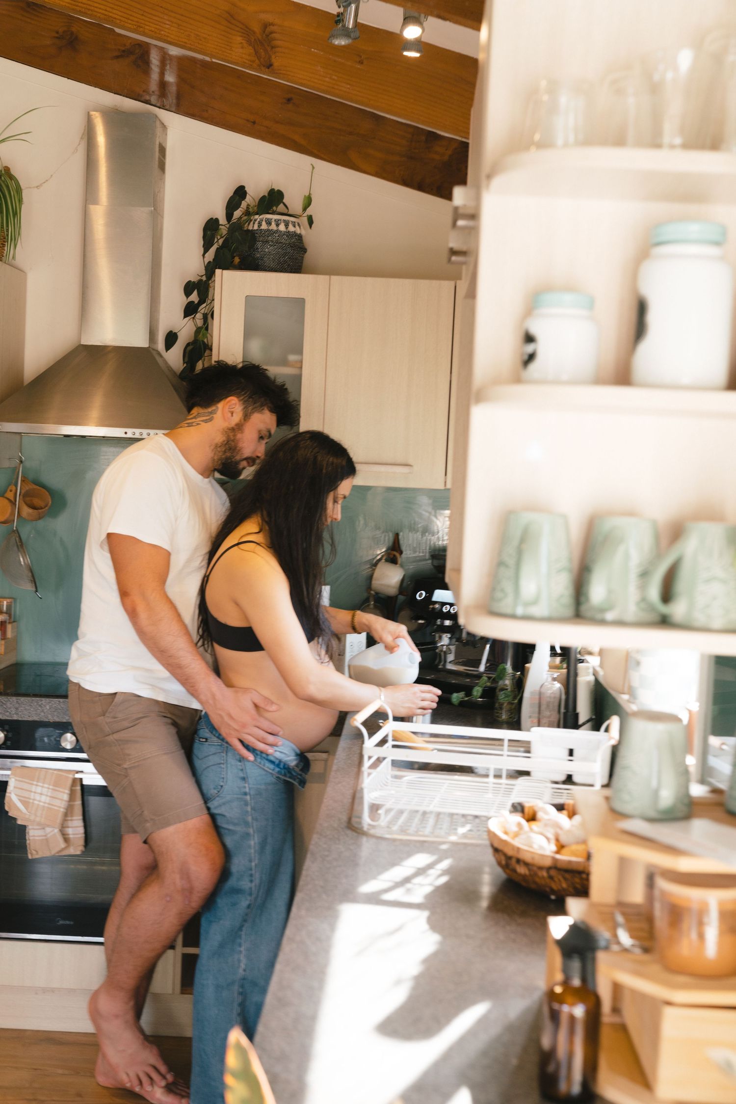 A couple shares a tender moment while cooking together in a sunlit kitchen with mason jars on floating shelves.